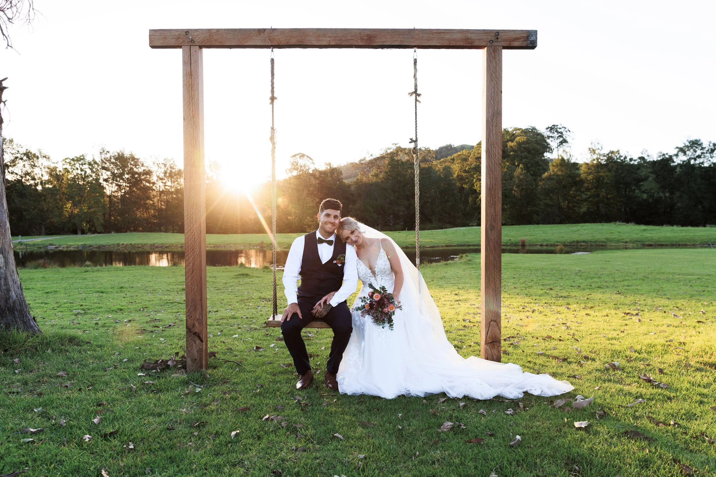 A newlywed couple sitting on a swing under a wooden frame outdoors during sunset. The groom is dressed in a black vest and bow tie, and the bride wears a white wedding gown and veil, holding a bouquet of flowers. Trees and a pond are visible in the b