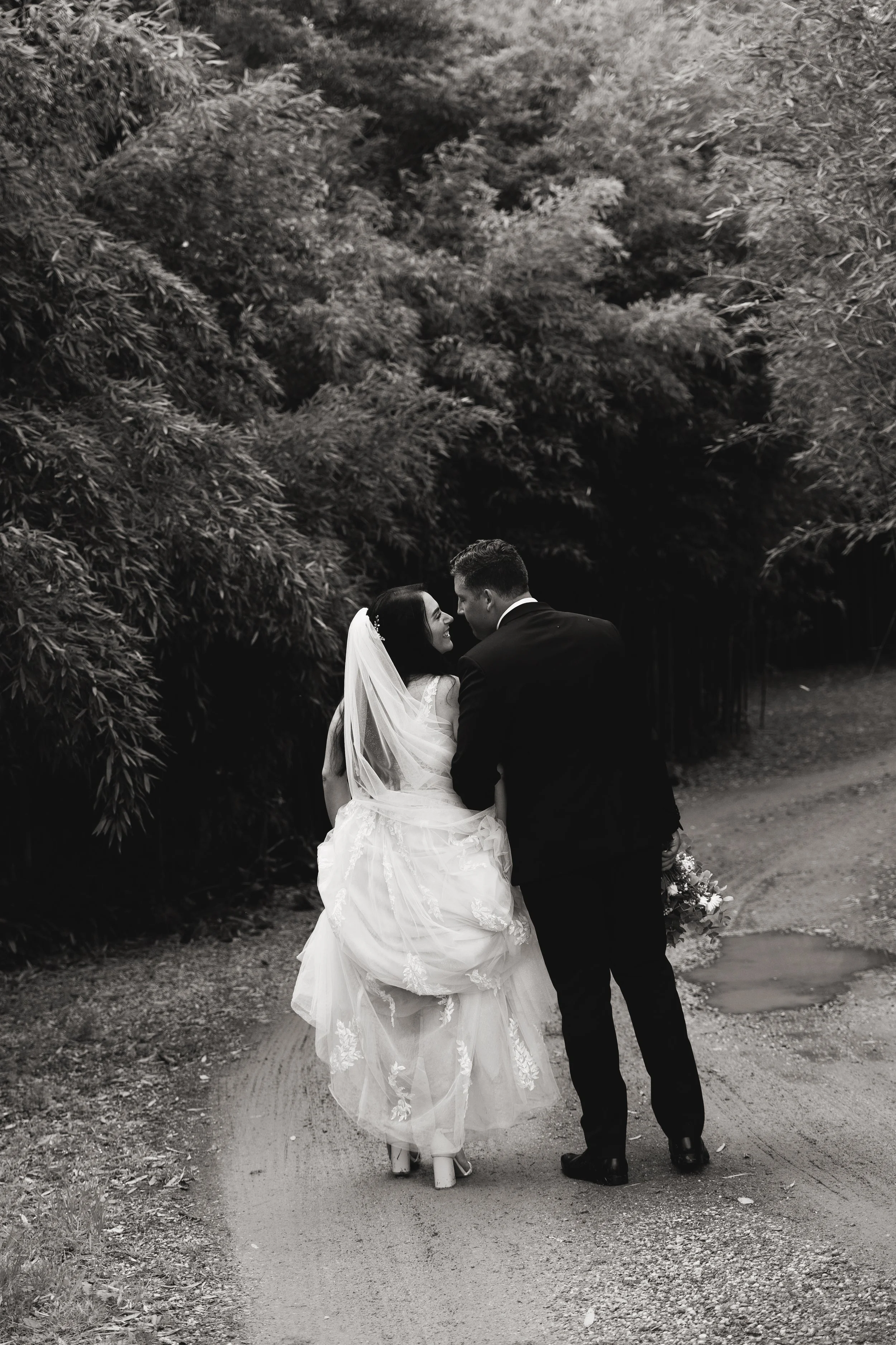Black and white photo of a newlywed couple walking on a dirt path, surrounded by large trees and bushes. The bride is wearing a white wedding dress with lace details and a veil, and the groom is in a dark suit. They are holding hands and looking at e