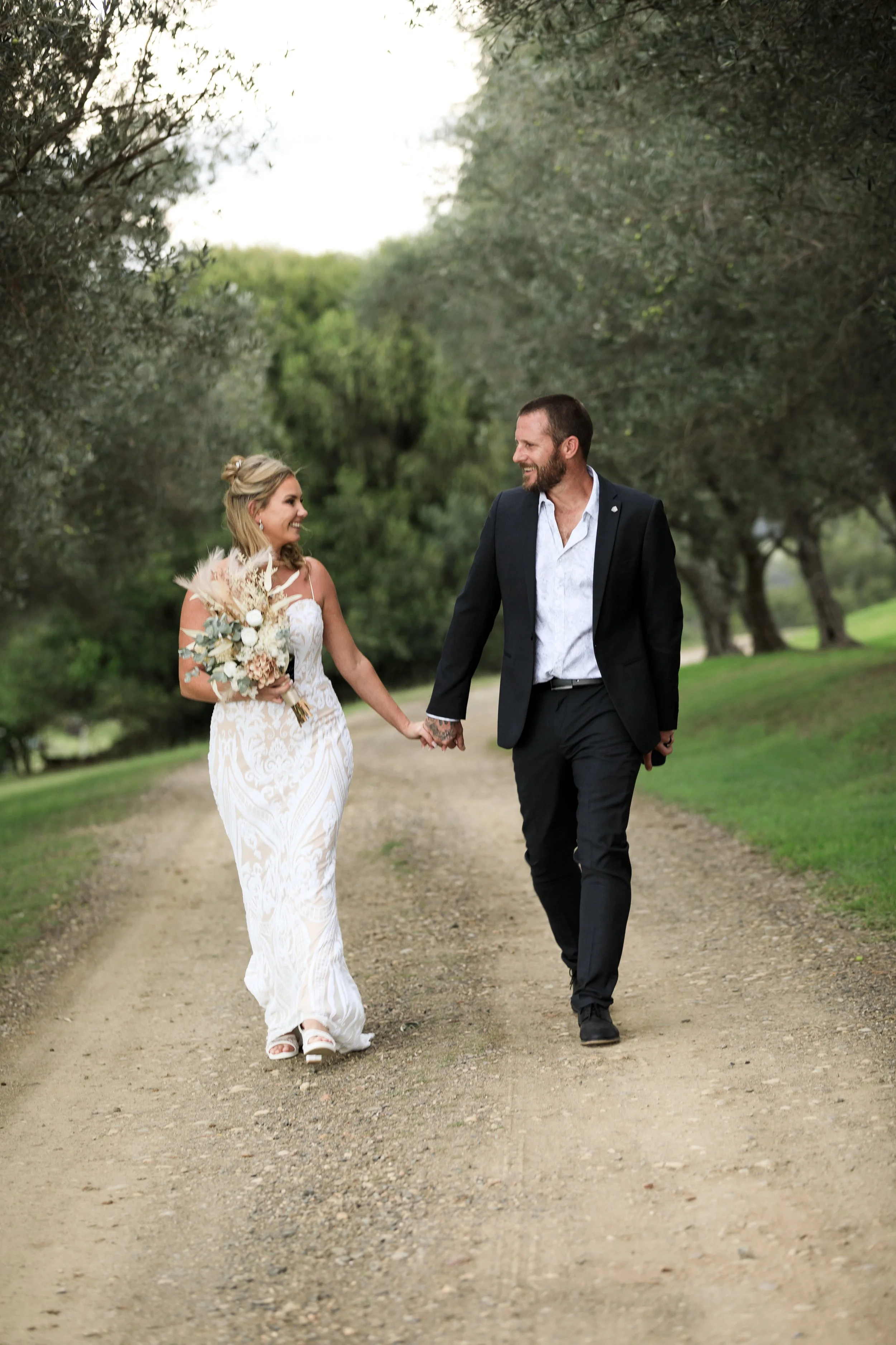 A newlywed couple walking hand in hand on a dirt path surrounded by green trees, smiling and looking at each other.