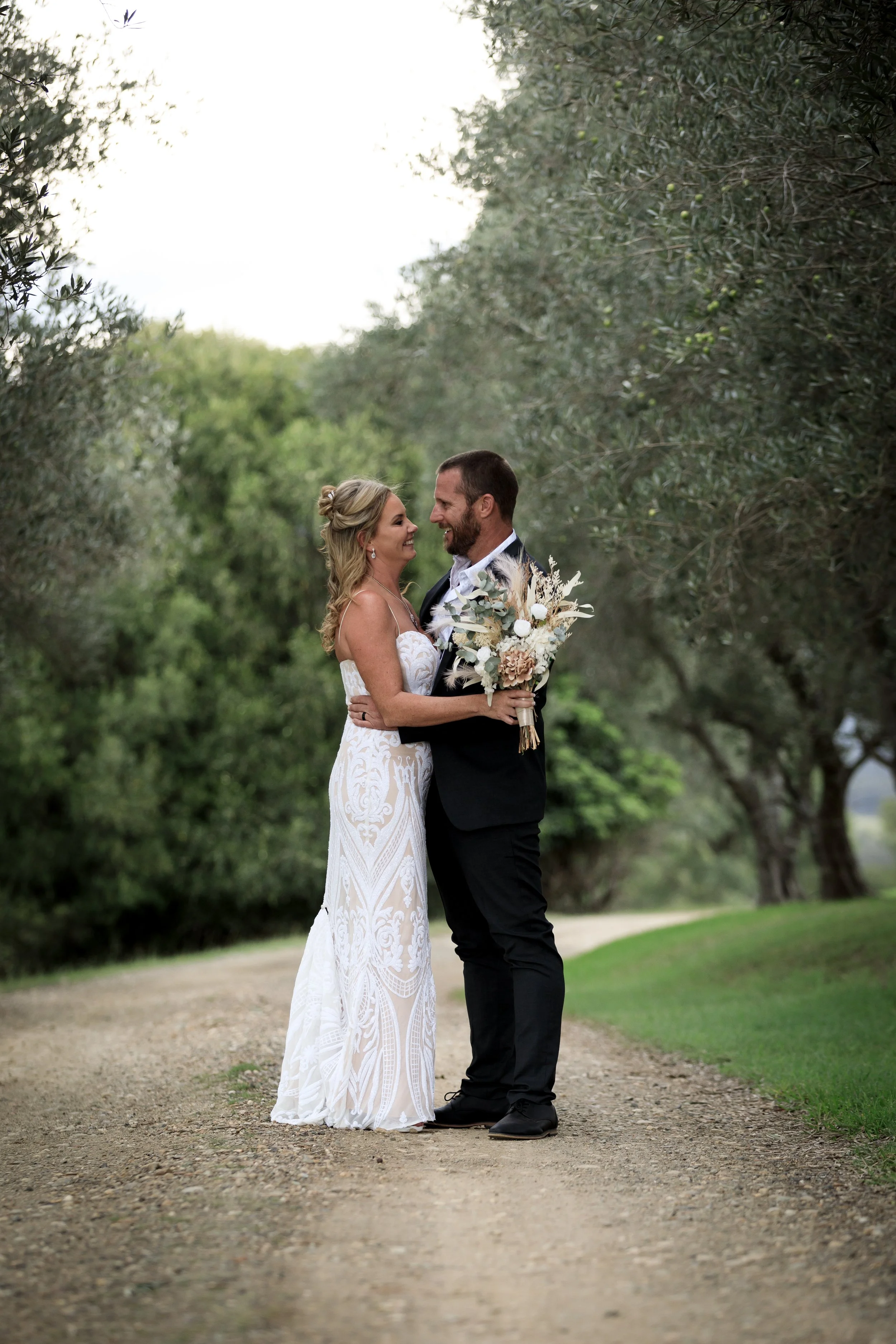A bride and groom standing outdoors on a dirt path, gazing at each other and smiling, surrounded by lush green trees, with the bride holding a bouquet of dried and white flowers.