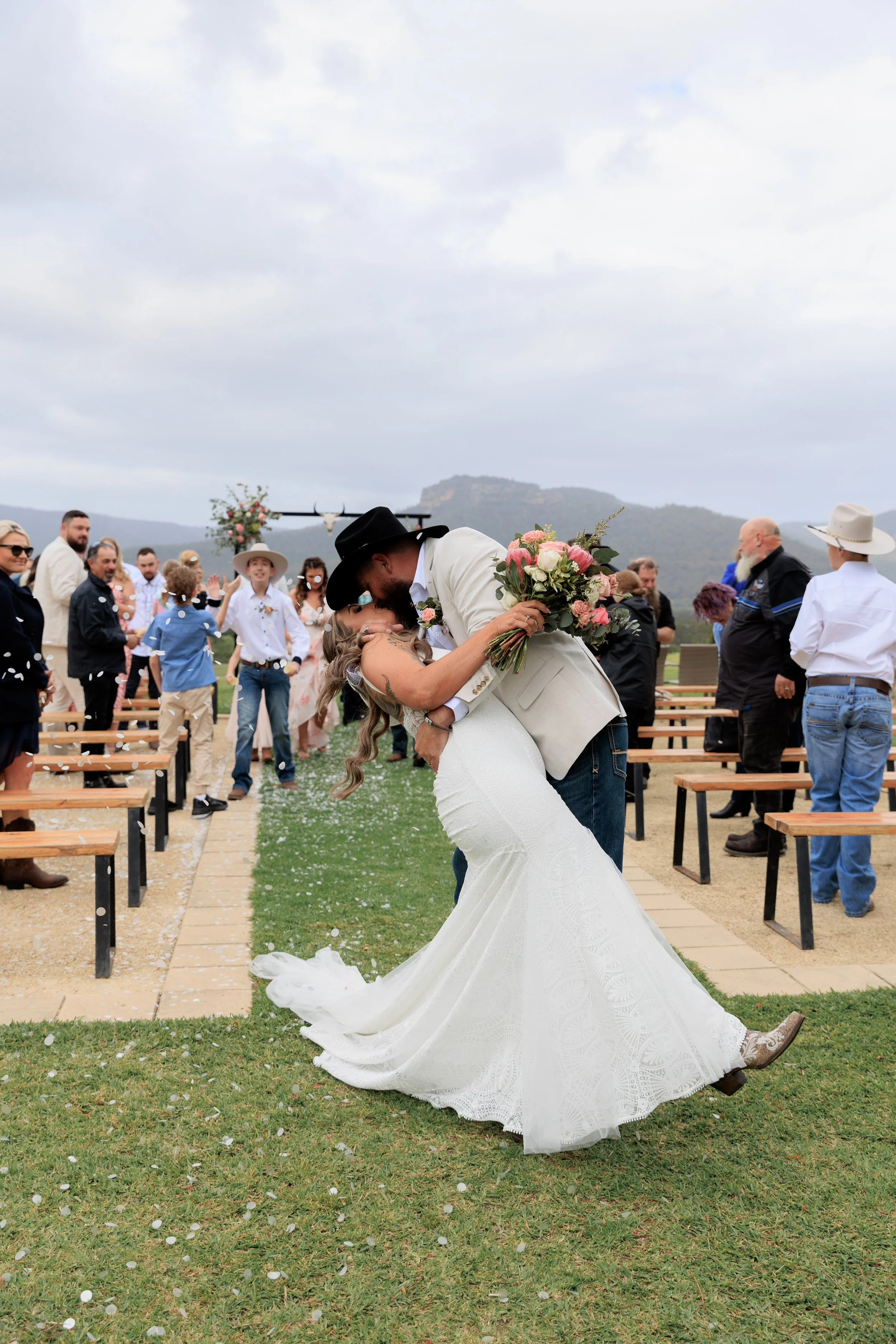 A newlywed couple sharing a kiss, with the bride holding a bouquet, surrounded by guests at an outdoor wedding ceremony in a scenic mountainous area.
