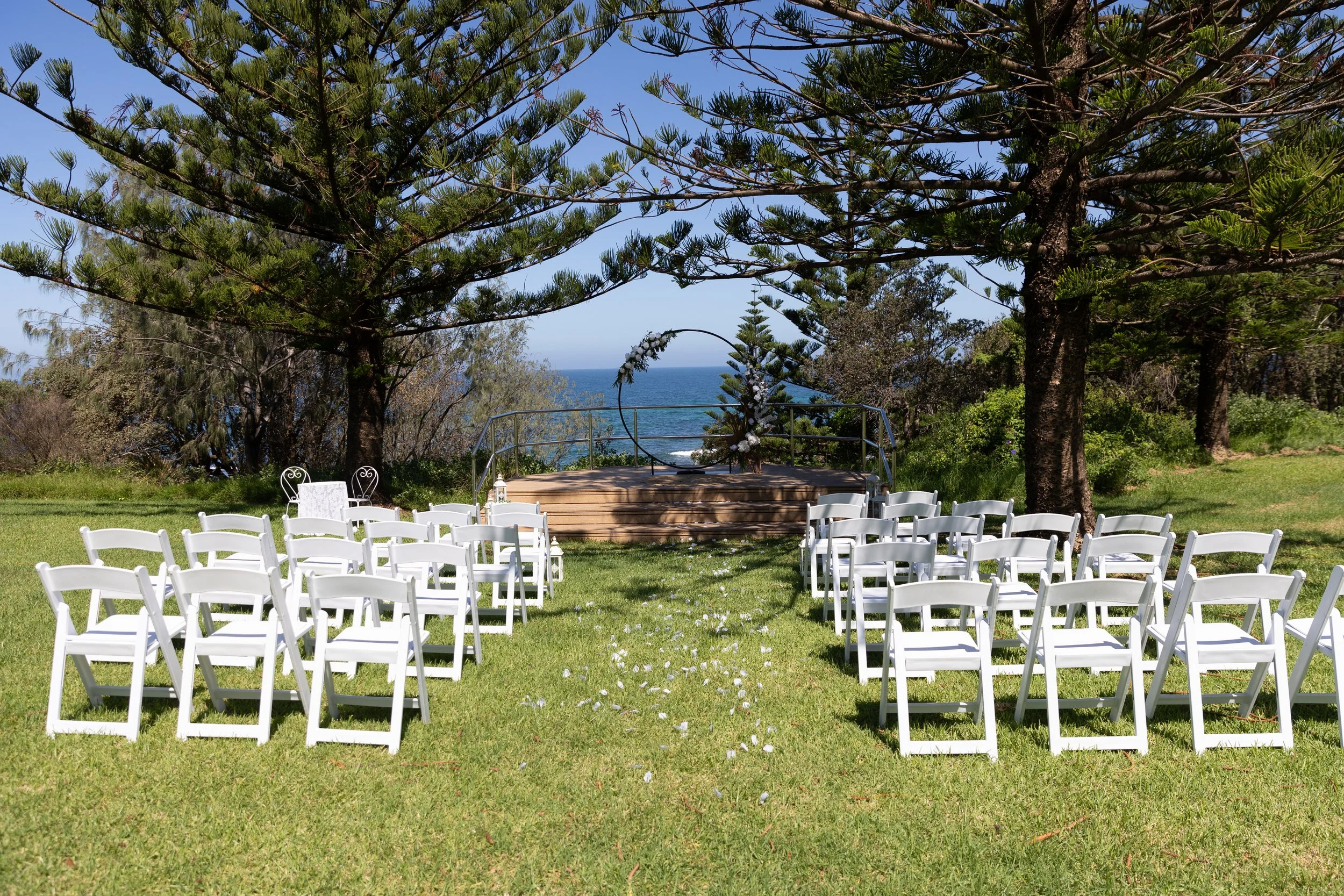Outdoor wedding setup with white chairs arranged on grass facing a small stage decorated with flowers, under large trees, overlooking the ocean.
