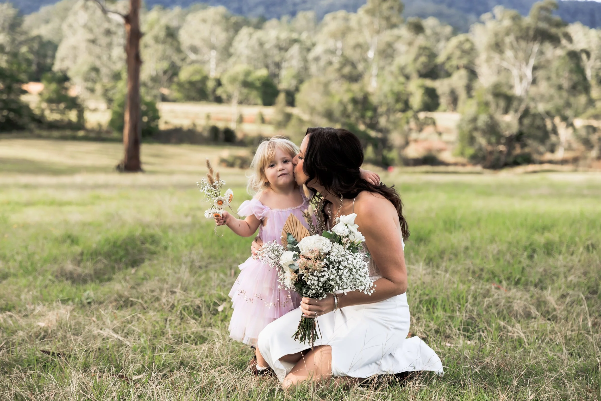 A woman dressed in white kneeling in a grassy field and kissing a young girl on her cheek. The girl has blonde hair, is wearing a pink dress, and holding a bouquet of flowers. The background features trees and a hilly landscape.