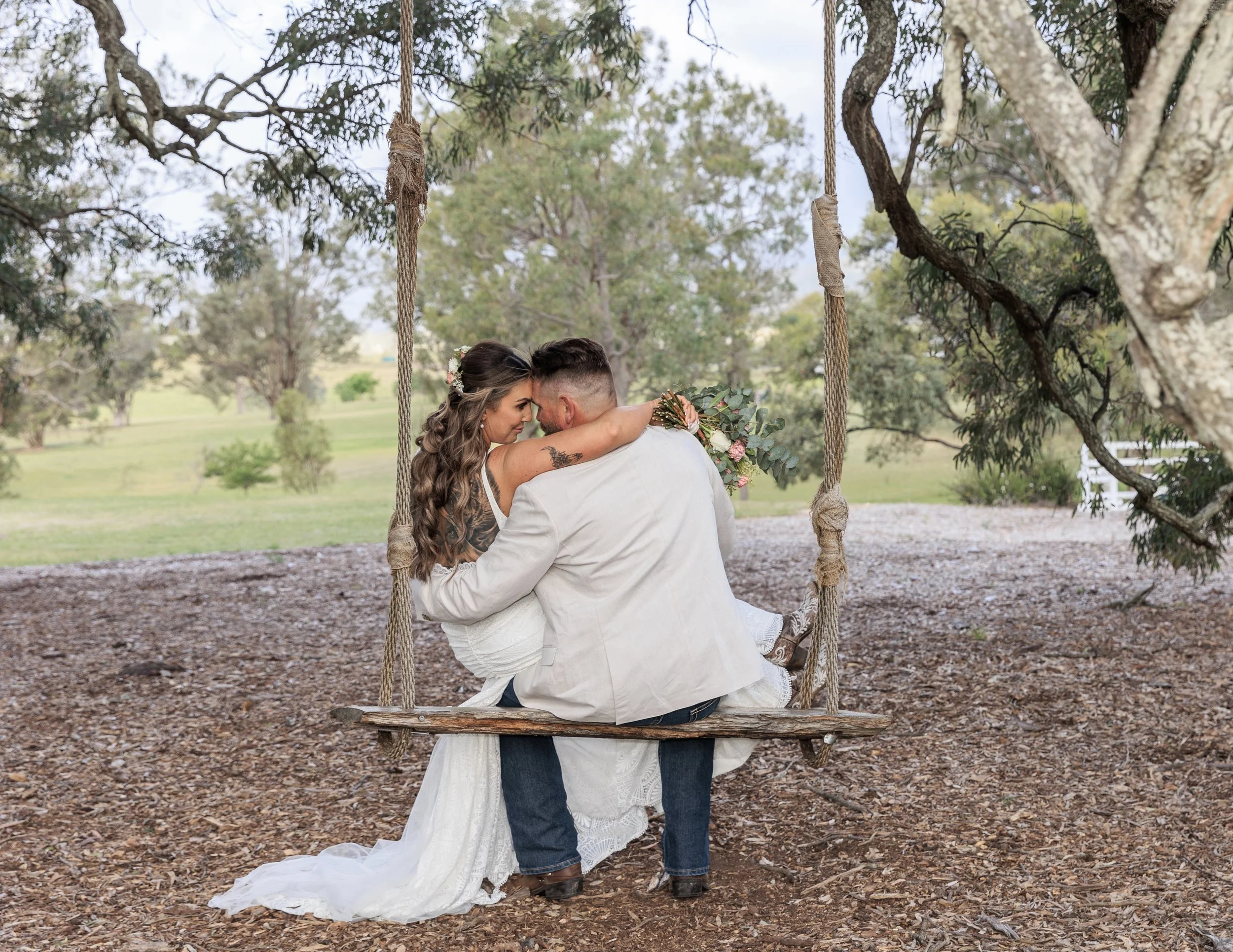 A couple sitting on a wooden swing in a park, embracing closely. The woman is wearing a white wedding dress and floral crown, and the man is in a white shirt and jeans. They are touching foreheads and holding a small bouquet.