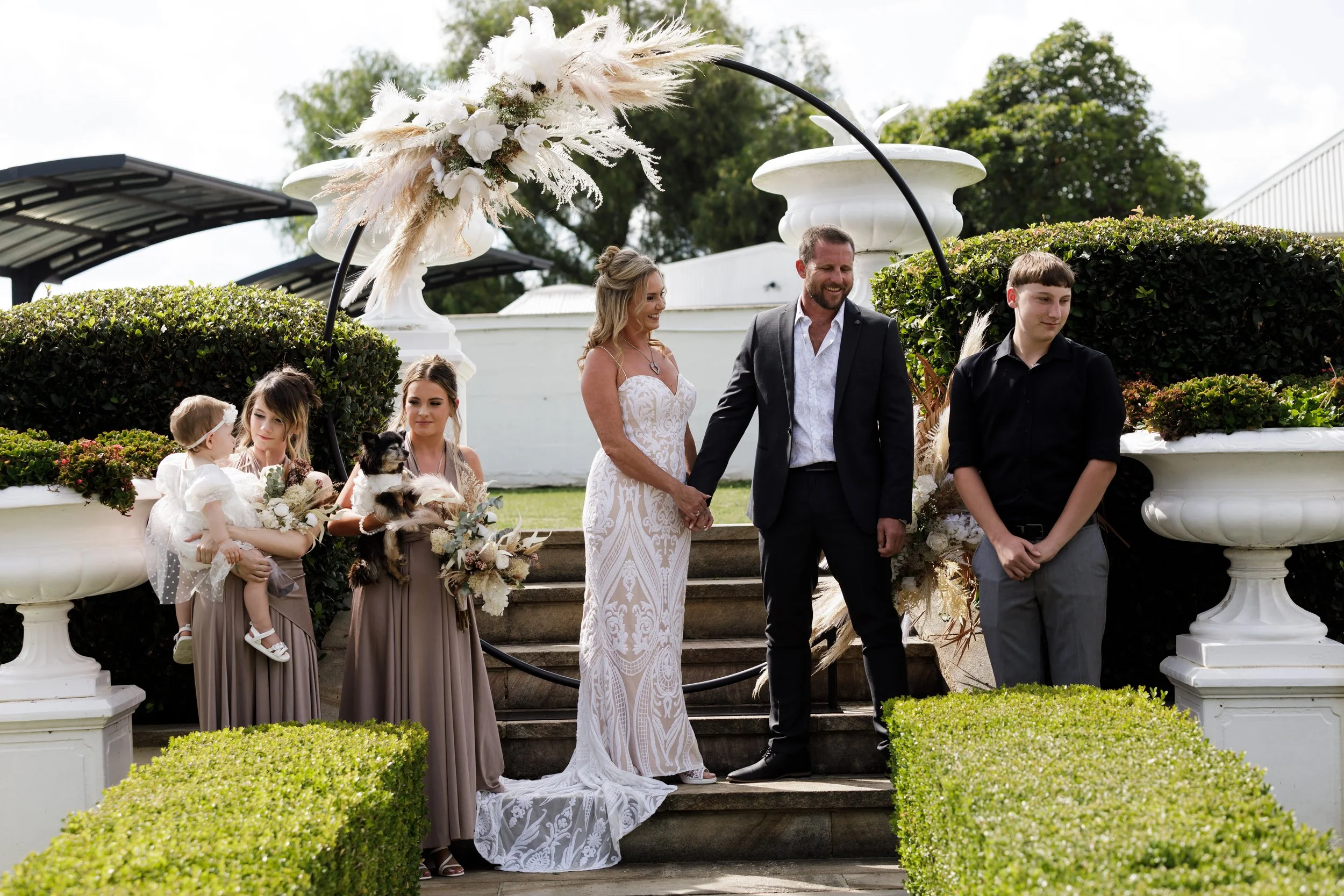 A wedding ceremony outdoors featuring a bride and groom holding hands on steps, surrounded by bridesmaids, flower girls, and greenery, with decorative floral arrangements overhead.