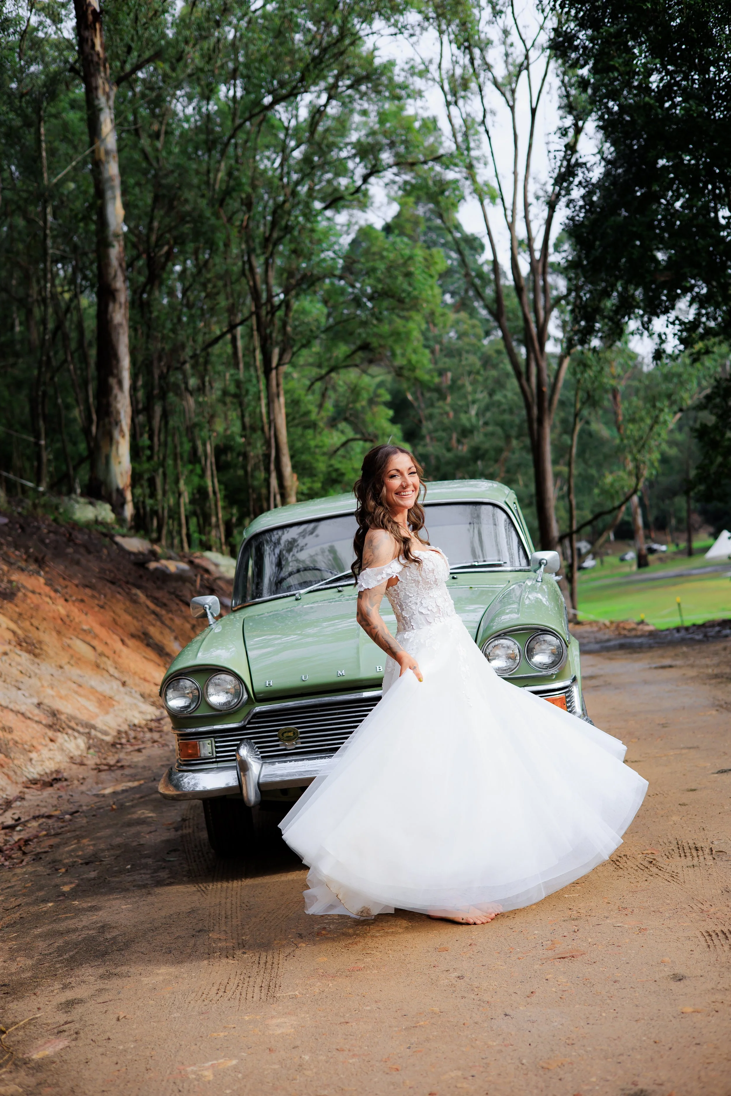 A woman in a white wedding dress standing in front of a green vintage Cadillac on a dirt path in a wooded area, smiling and looking at the camera.