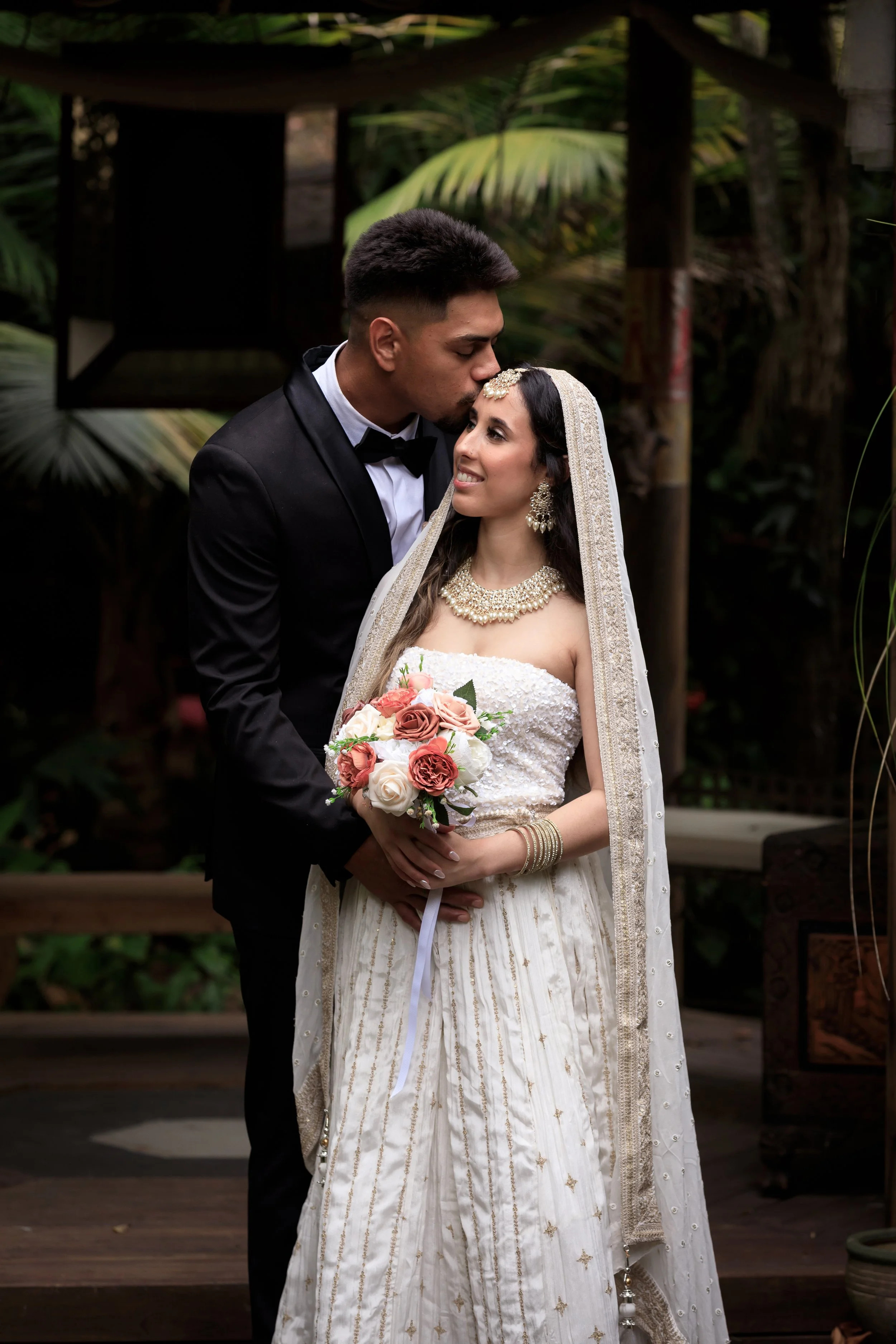 A groom in a black tuxedo kisses a bride in a white wedding dress holding a bouquet of pink and white roses. They stand outdoors surrounded by green foliage.