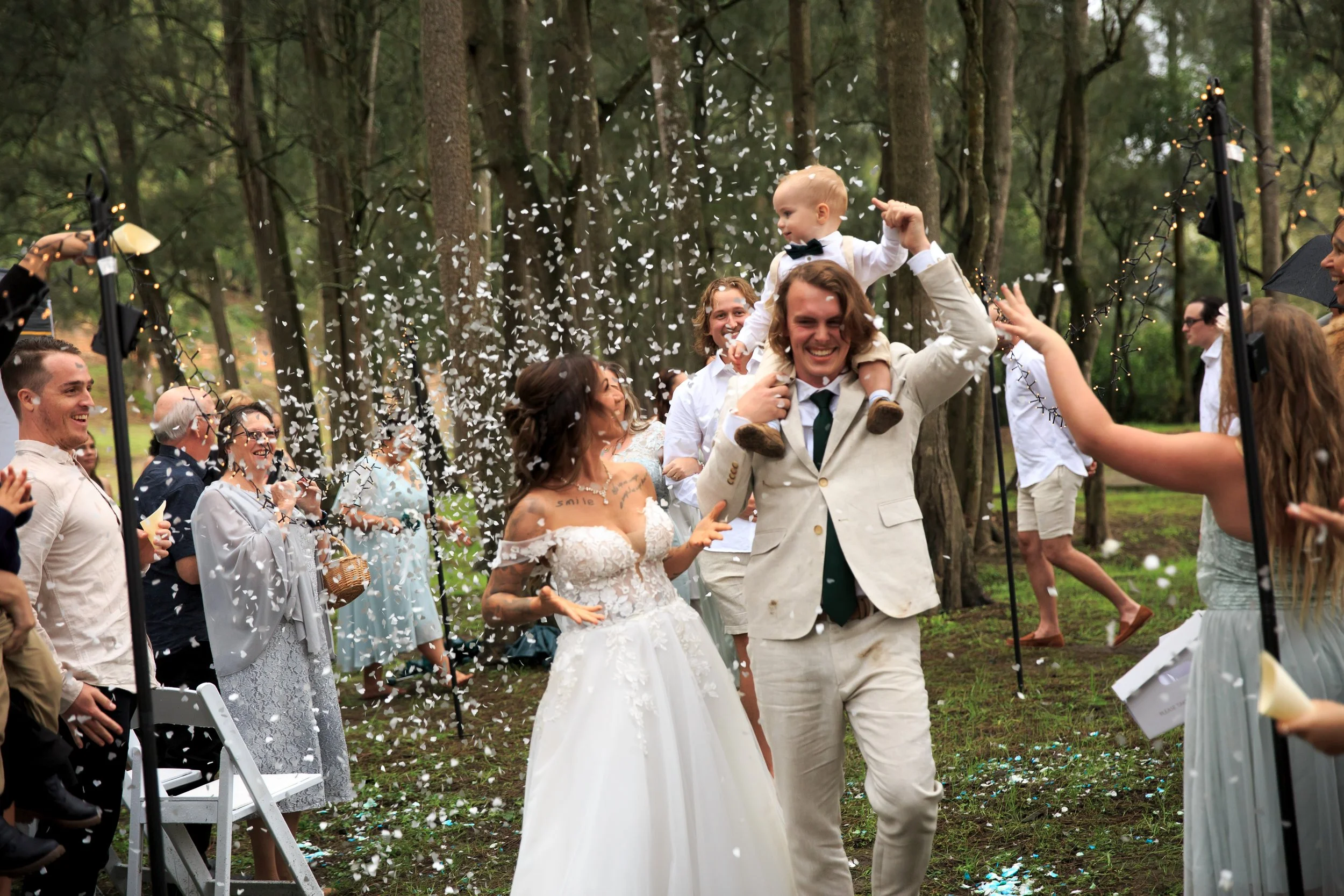 A wedding celebration outdoors in a wooded area with a group of people throwing confetti. A man in a beige suit is holding a young child on his shoulders, both smiling. Guests are clapping and cheering around them.