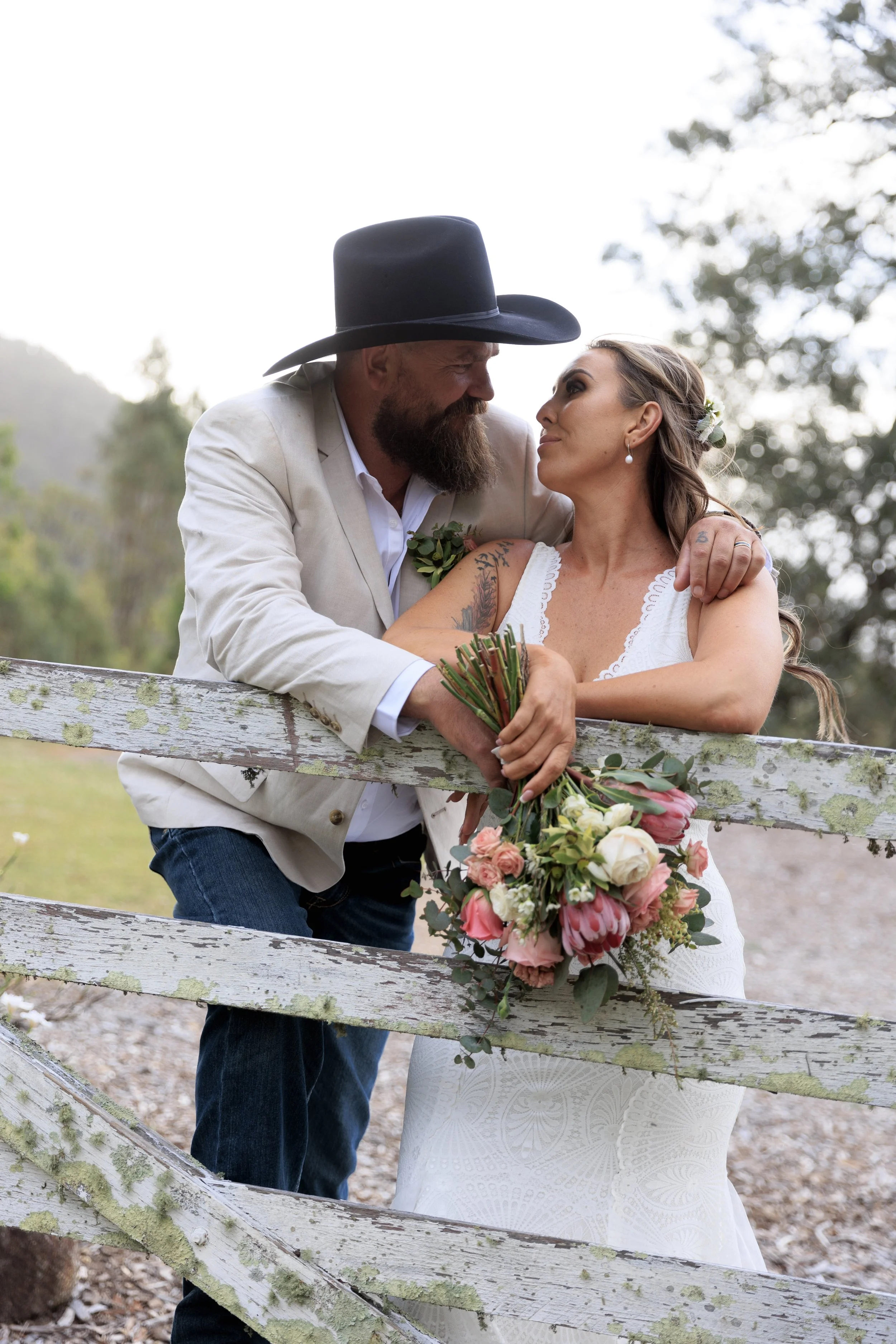 A couple on a wooden fence, dressed in white wedding clothes, gazing at each other. The man wears a cowboy hat and has a beard, while the woman has wavy hair and floral accessories. She holds a large bouquet of pink and white flowers. The background 