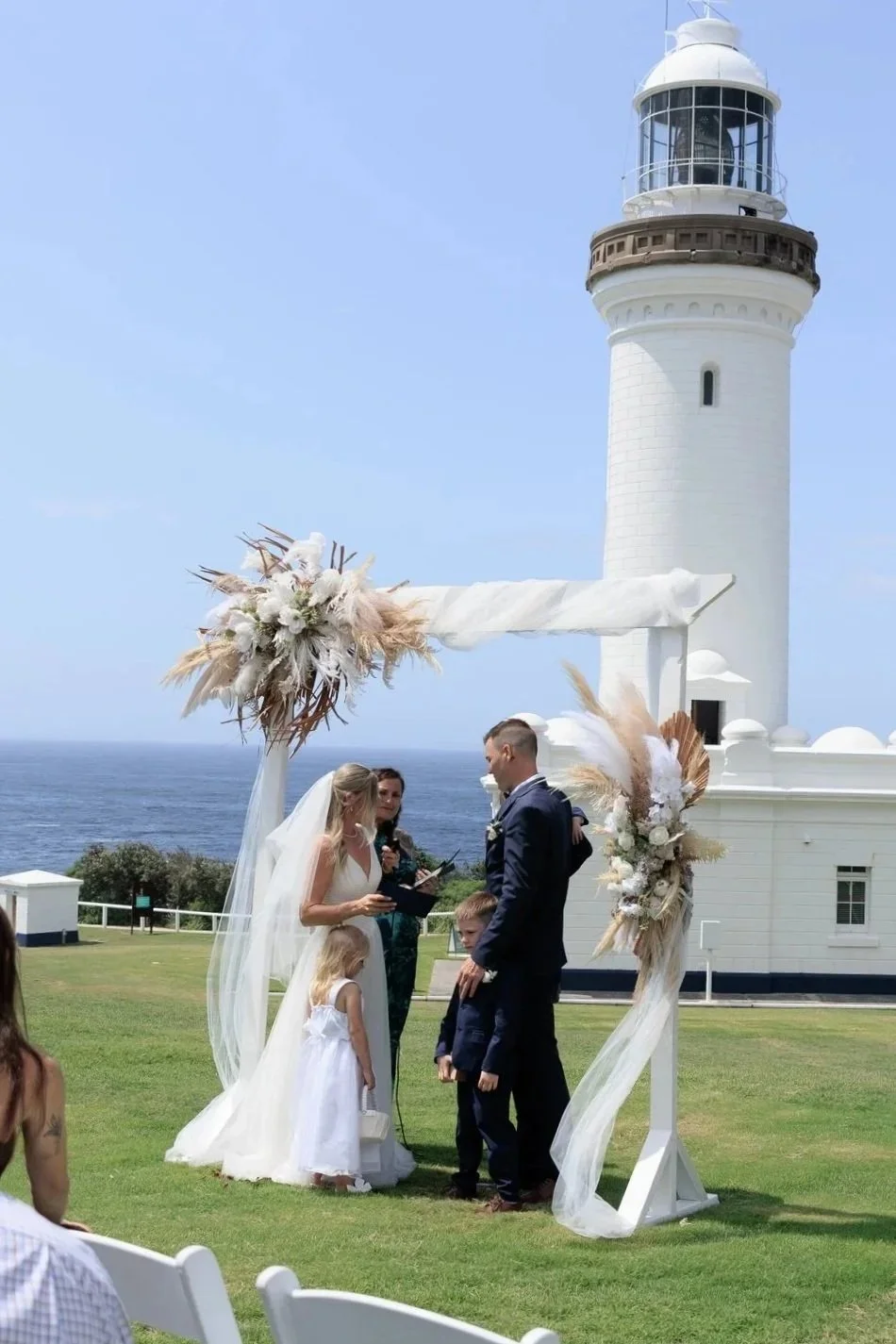 A wedding ceremony taking place outdoors next to a white lighthouse by the ocean. The bride and groom are standing under a decorated arch with floral arrangements and sheer fabric. The bride is wearing a white wedding dress and veil, the groom a dark suit. Two children, a girl in a white dress and a boy in a suit, are also present.