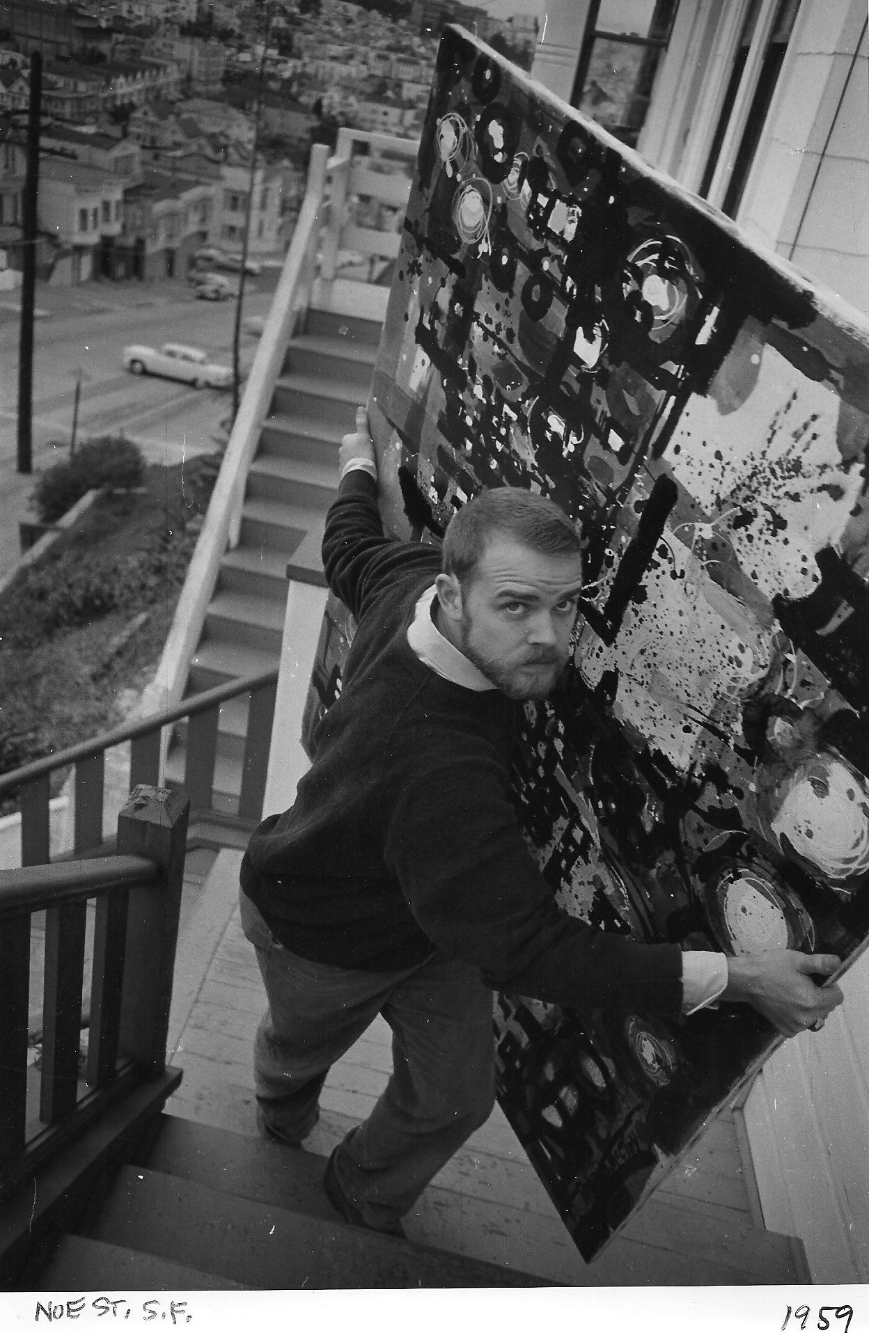 McClay looking up at the camera while carrying his painting, "Night City" on the staircase of his San Francisco apartment, with cars and houses visible in the background.
