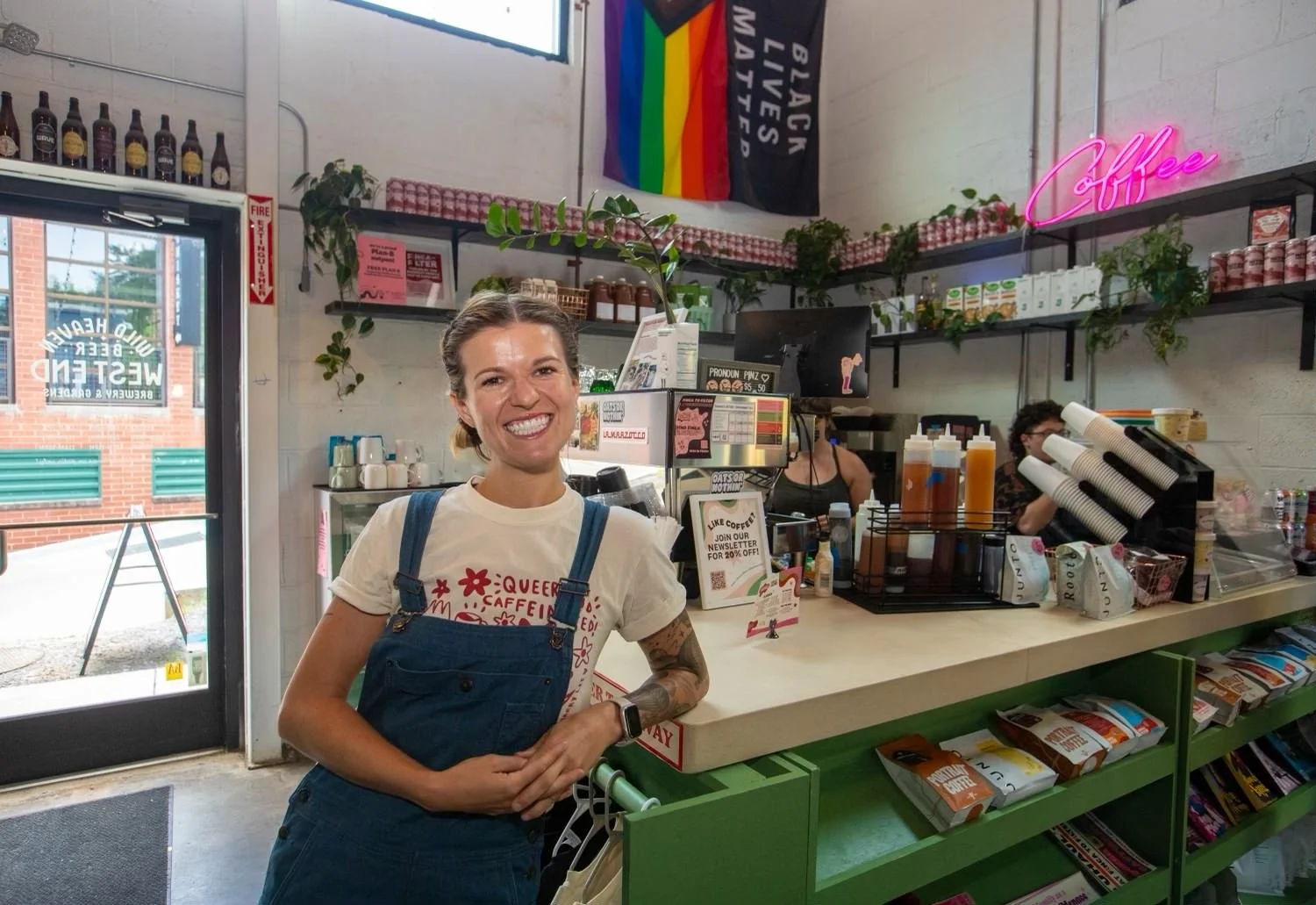 Smiling woman in a white queer-themed T-shirt and denim overalls standing behind the counter of a coffee shop, with rainbow and pride flags on the wall and various coffee supplies around.