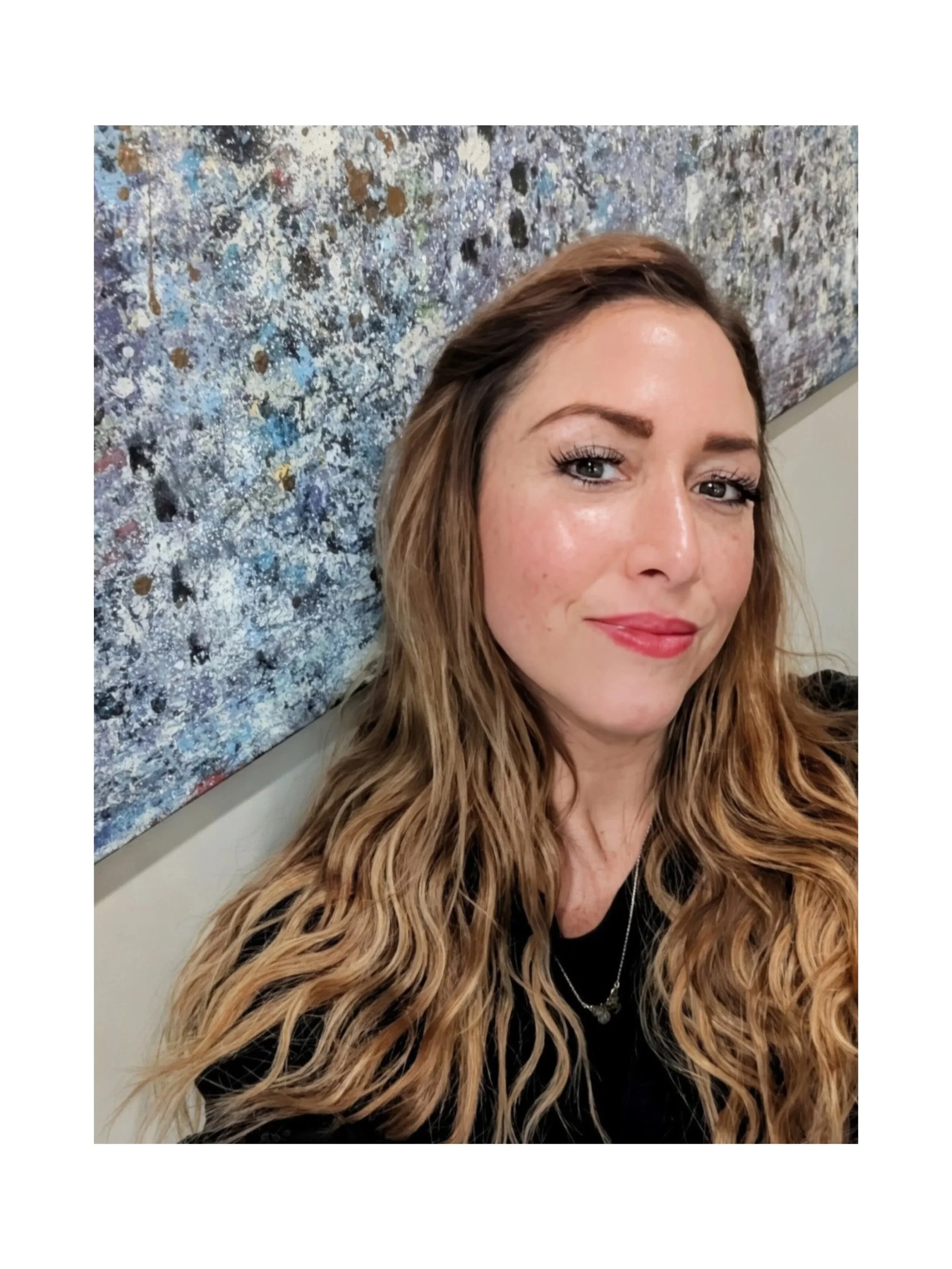 Close-up selfie of a woman with long wavy blonde hair, wearing a black top and jewelry, standing in front of an abstract painting with splashes of blue, white, and brown.