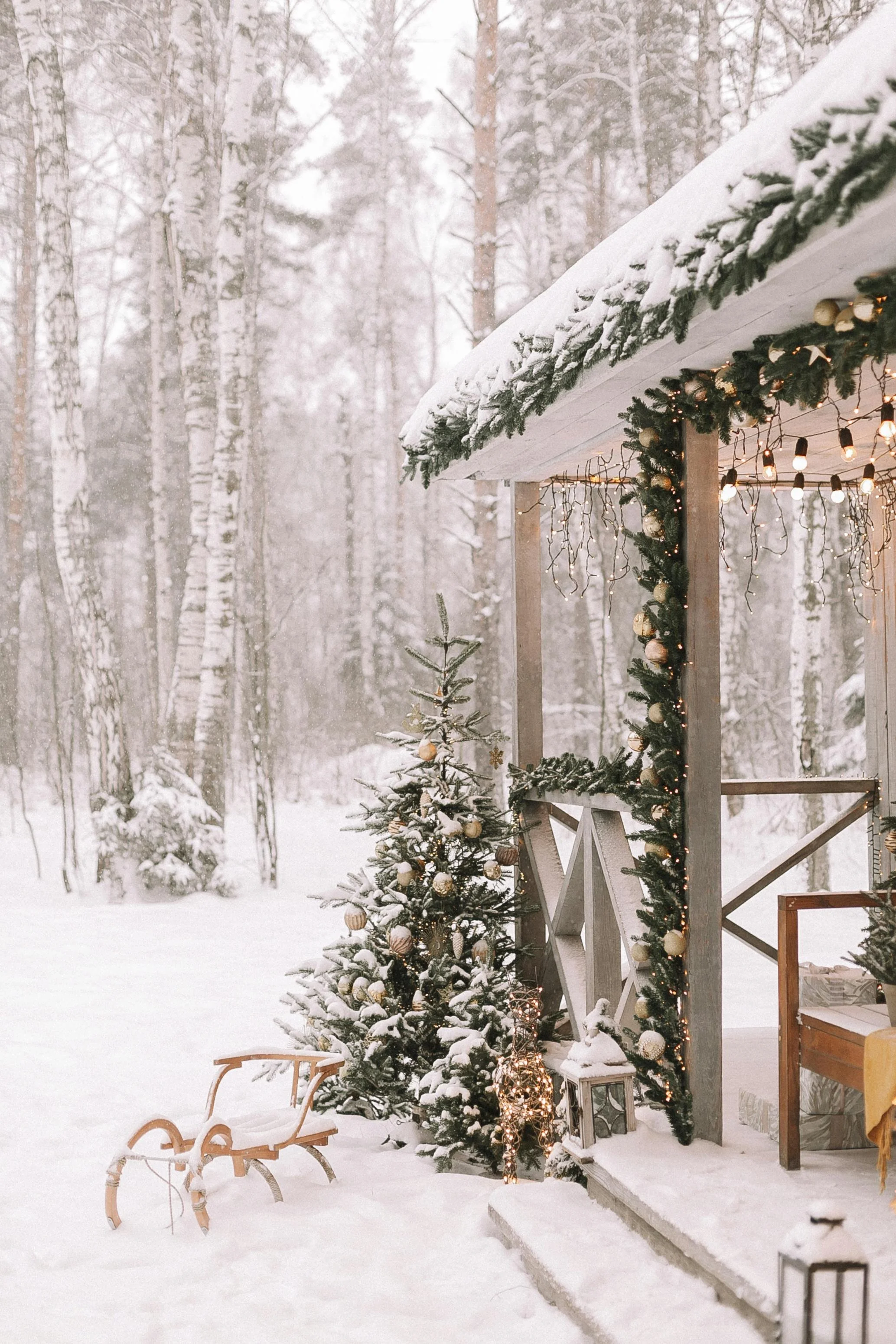 Snow-covered Christmas decorations on an outdoor porch, including a decorated Christmas tree, string lights, and garland, with a snowy forest in the background.