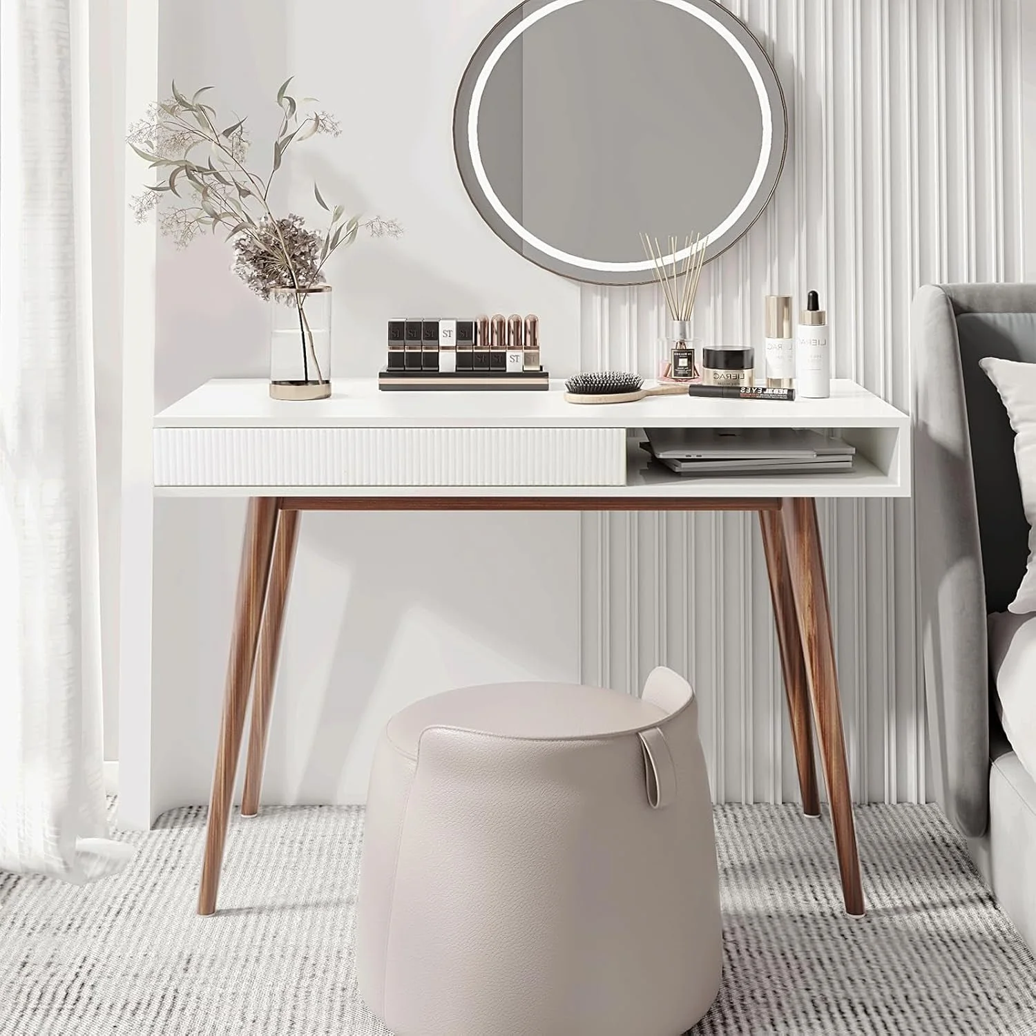 A white vanity table with wooden legs, situated in front of a white textured wall and a mirror with LED lights. The table has various cosmetic products, a dried flower arrangement, a black hairbrush, and a reed diffuser. There is a white upholstered 