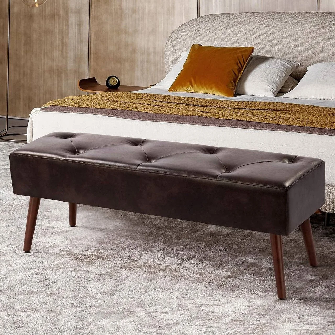 View of a black leather upholstered bench with wooden legs placed in front of a bed with decorative pillows and bedding, in a bedroom setting.