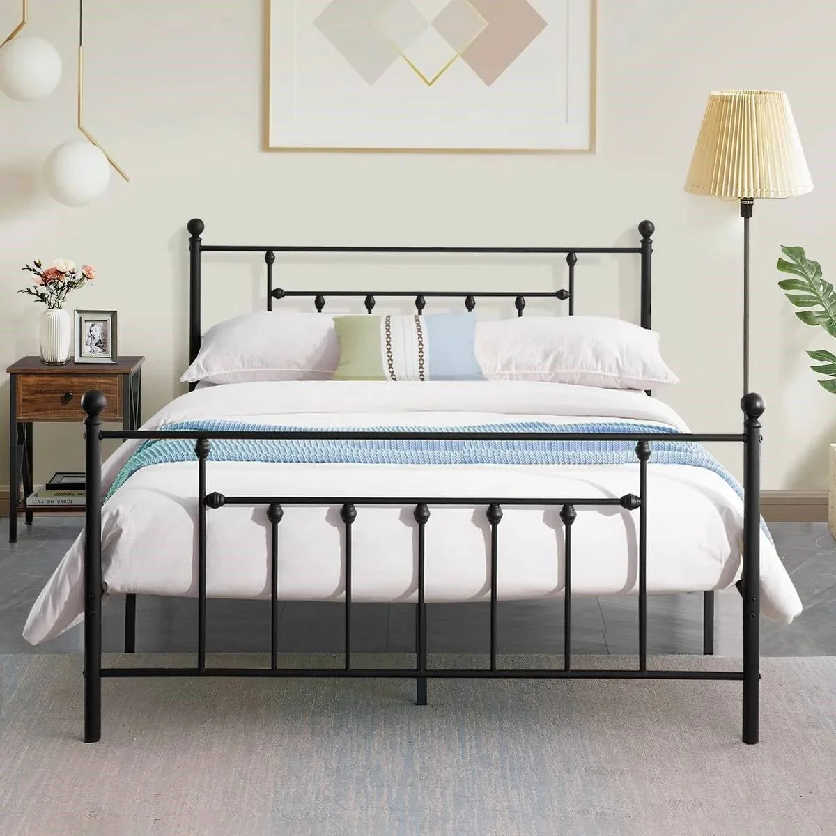 A bedroom with a black metal bed, white bedding, and a pillow with a blue and green stripe. There is a wooden nightstand with a vase of flowers, a framed photo, and a book. A floor lamp with a beige shade is on the right, and a green plant on the far
