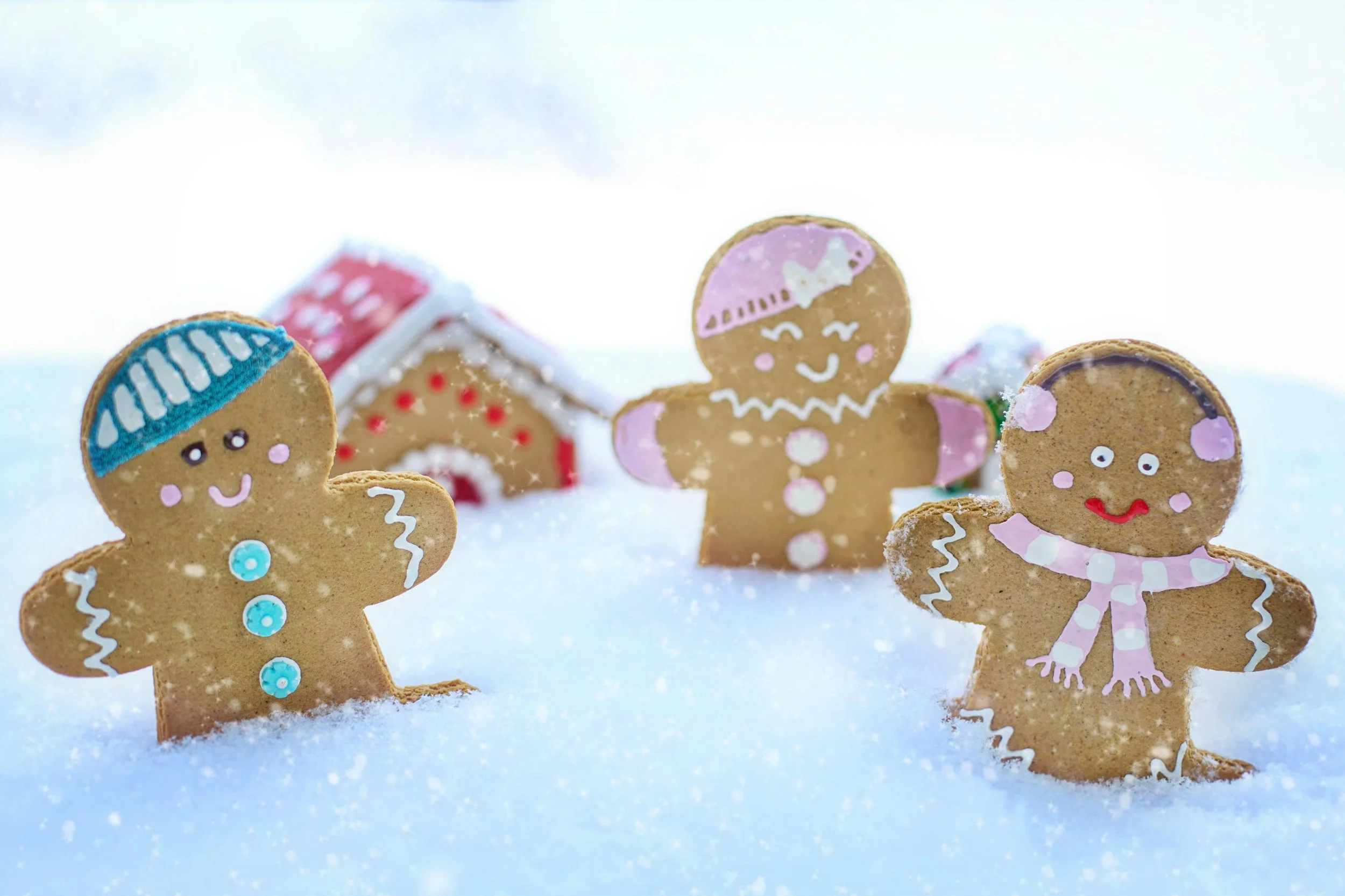 Decorated gingerbread cookies in snow with a small gingerbread house in the background.