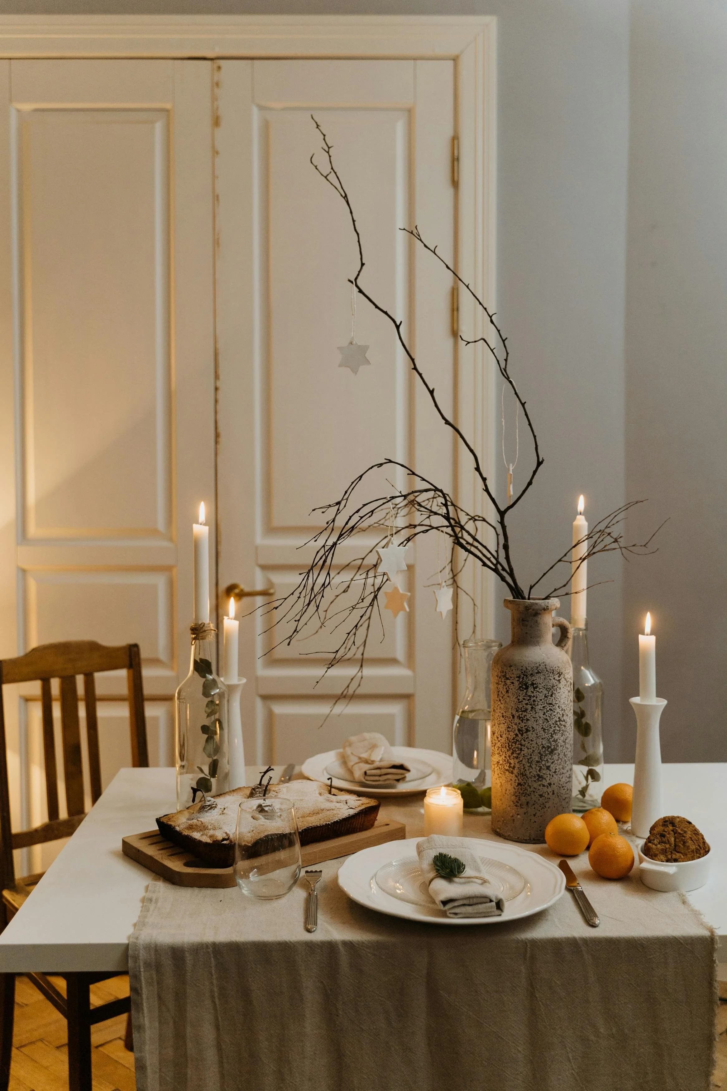 A cozy dining table set for a meal with white plates, cloth napkins, and cutlery. The table has a beige tablecloth, candles, and a large rustic vase with bare branches and hanging star ornaments as a centerpiece. There are oranges, a cake, and glass pitchers on the table, with warm candlelight creating a welcoming atmosphere.