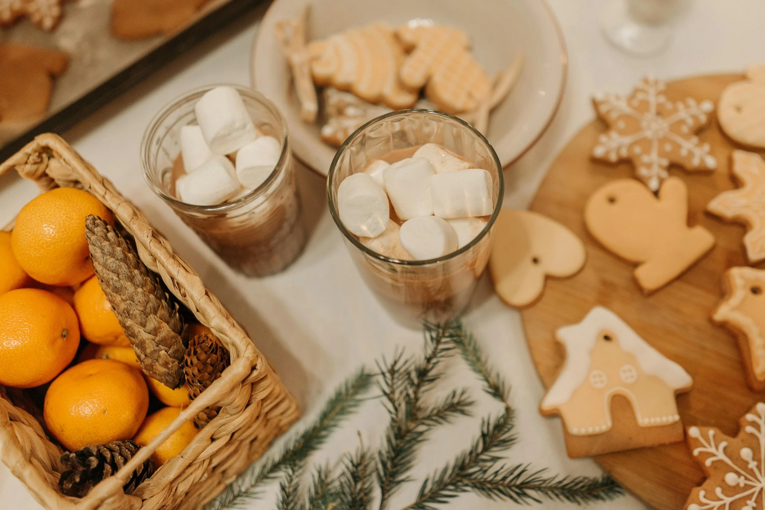 Two glasses of hot chocolate topped with marshmallows, a basket of oranges and pinecones, holiday cookies shaped like hearts, snowflakes, and gingerbread houses on a wooden board, greenery, and a white tablecloth.