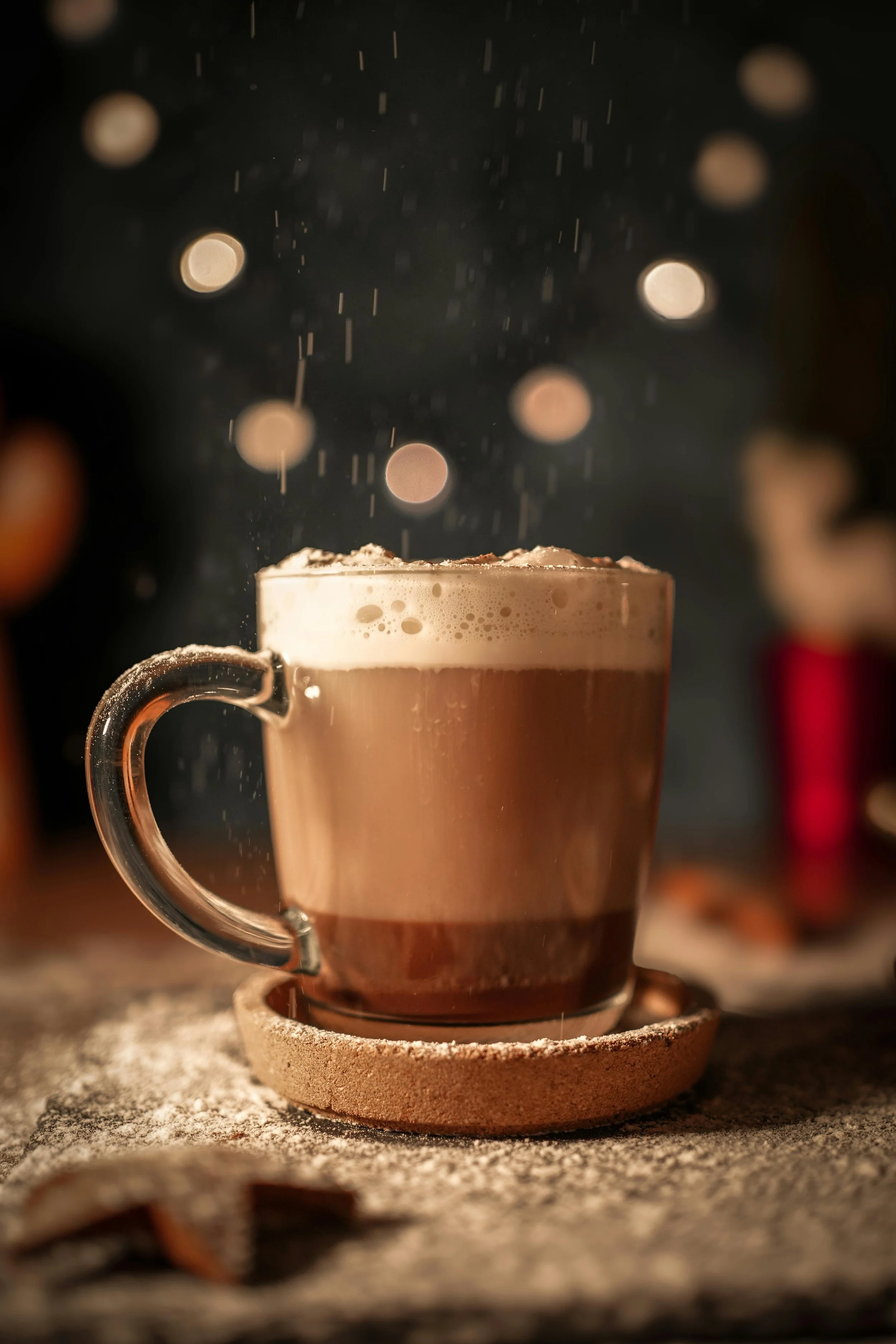 A glass mug filled with frothy hot chocolate or coffee, placed on a round cork coaster, with snow-like powder and falling snow or rain in the background.