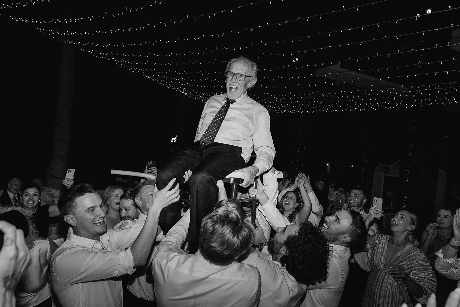 Grupo de personas celebrando en un evento, levantando a un hombre mayor que se encuentra sentado en una silla. Todos lucen felices y sonrientes, con decoración de luces en el techo.