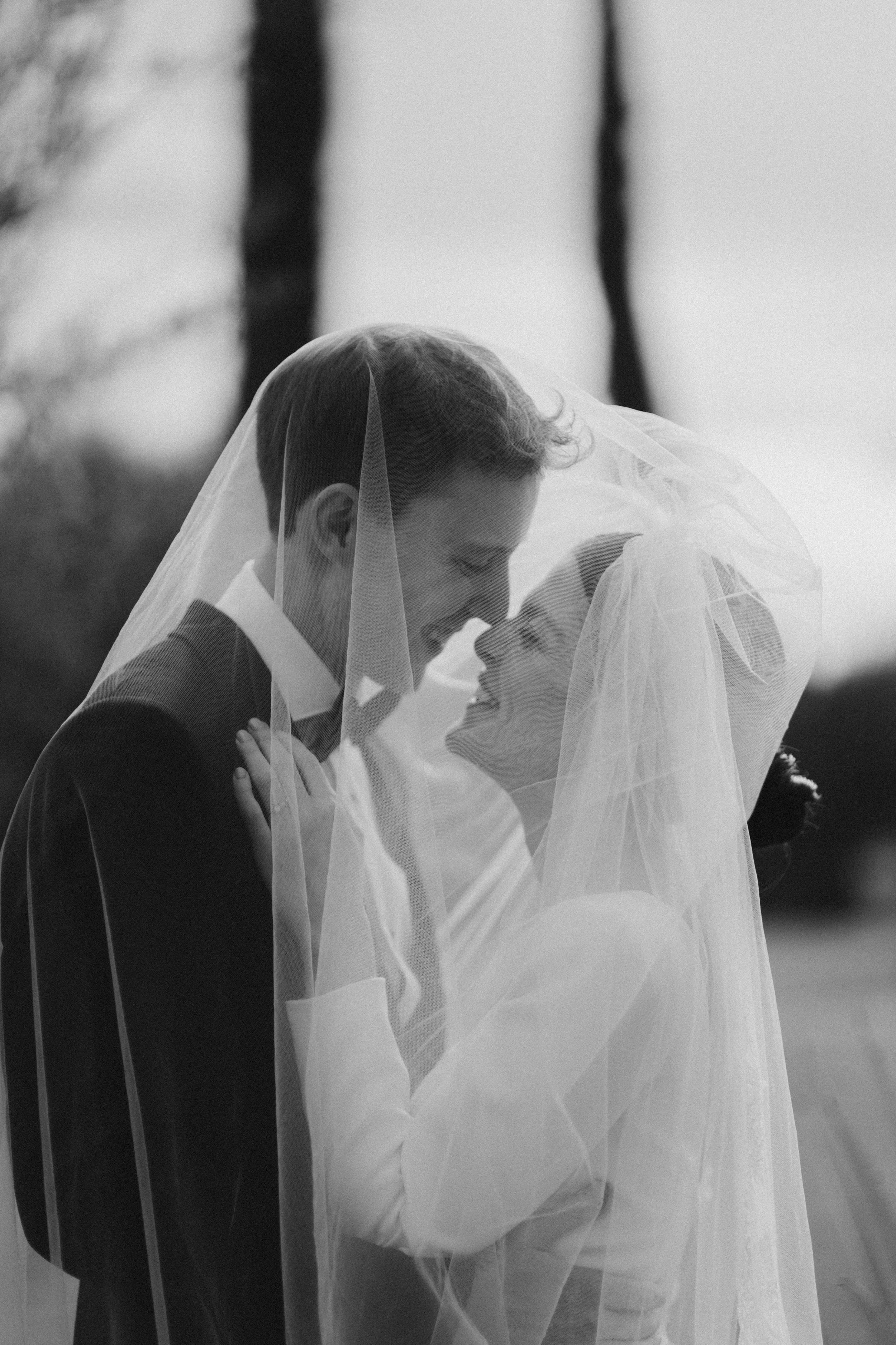 Una pareja de novios en una boda, cubiertos con un velo de novia, sonriendo y tocándose la cara mientras se miran con amor.