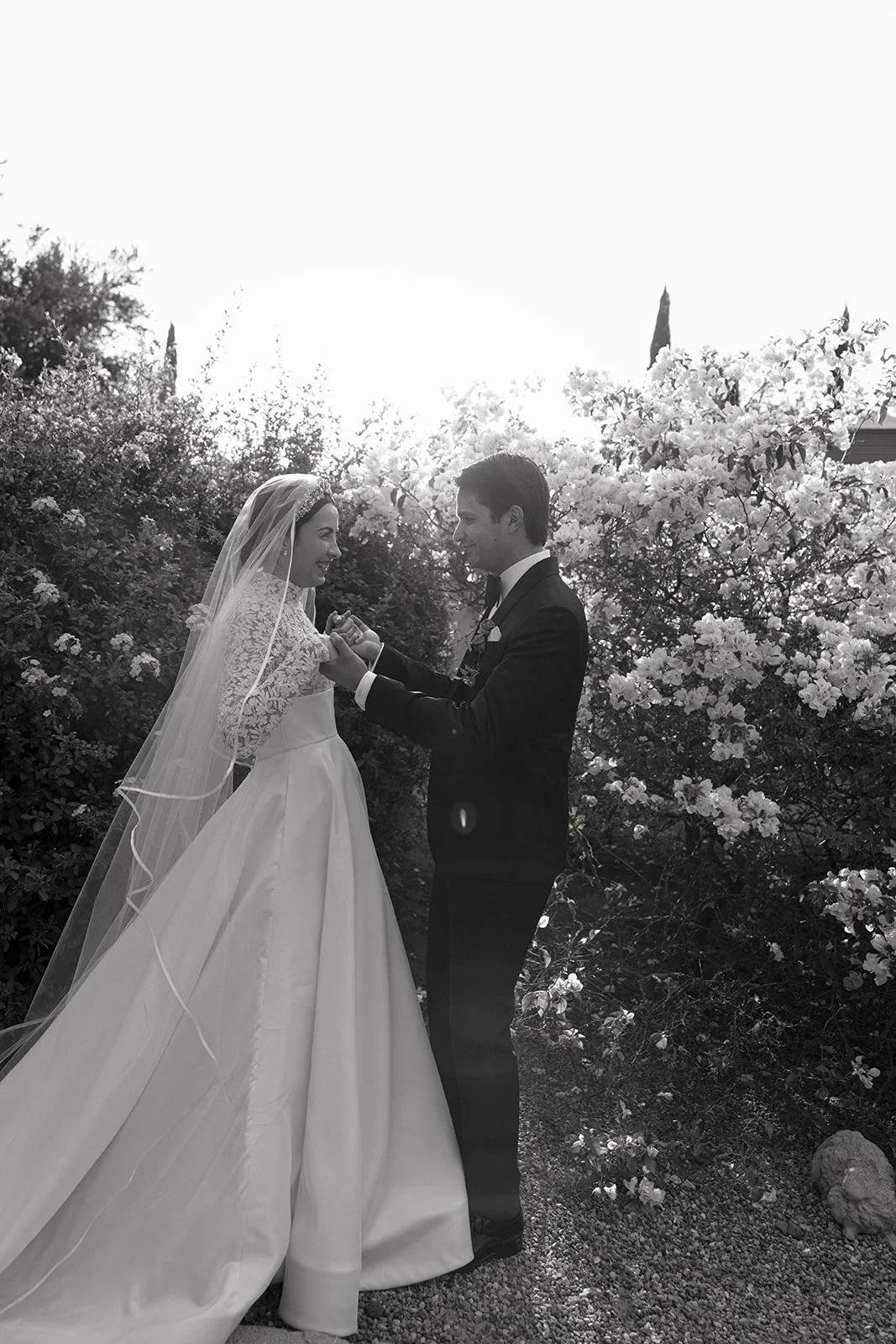 Pareja de novios en boda, la novia con vestido largo y velo, el novio con traje negro, en un jardín con flores, en un momento emotivo.