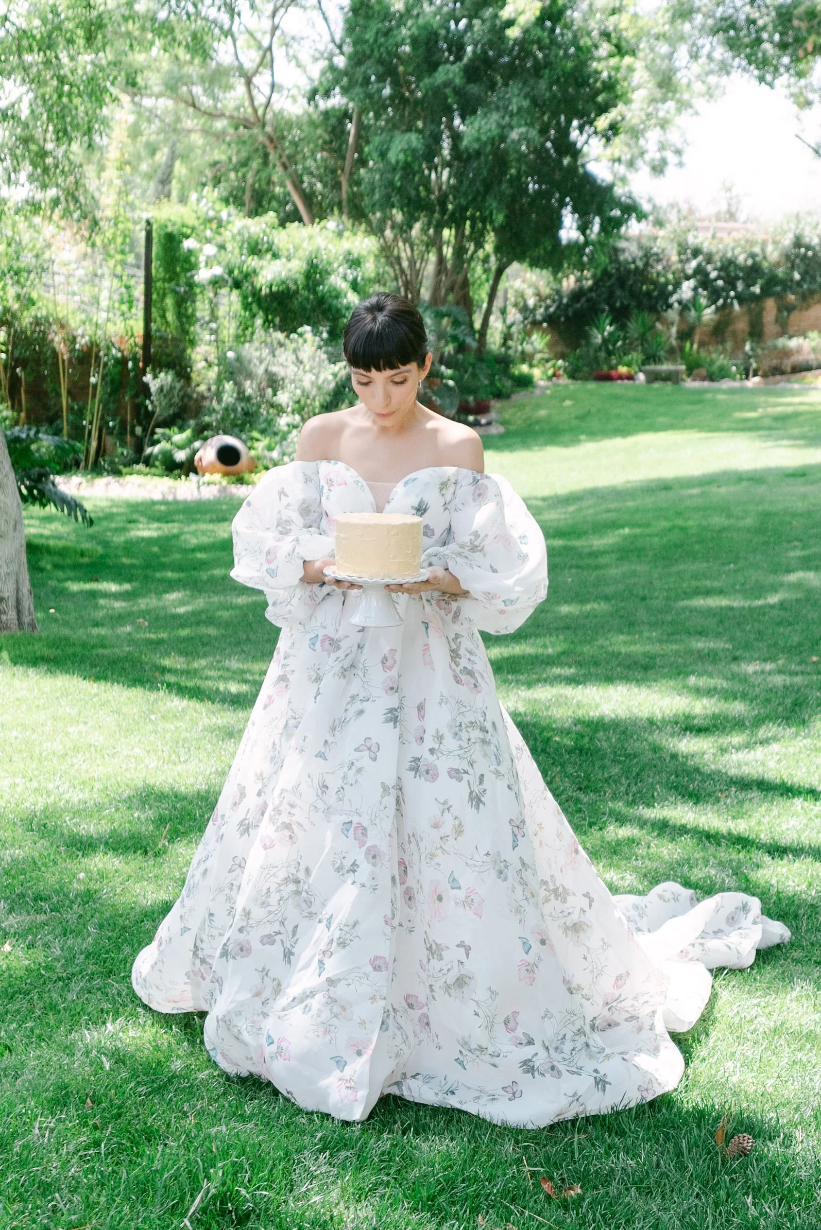 Una mujer con vestido largo y de manga larga sostiene un pastel en un jardín con césped y árboles.