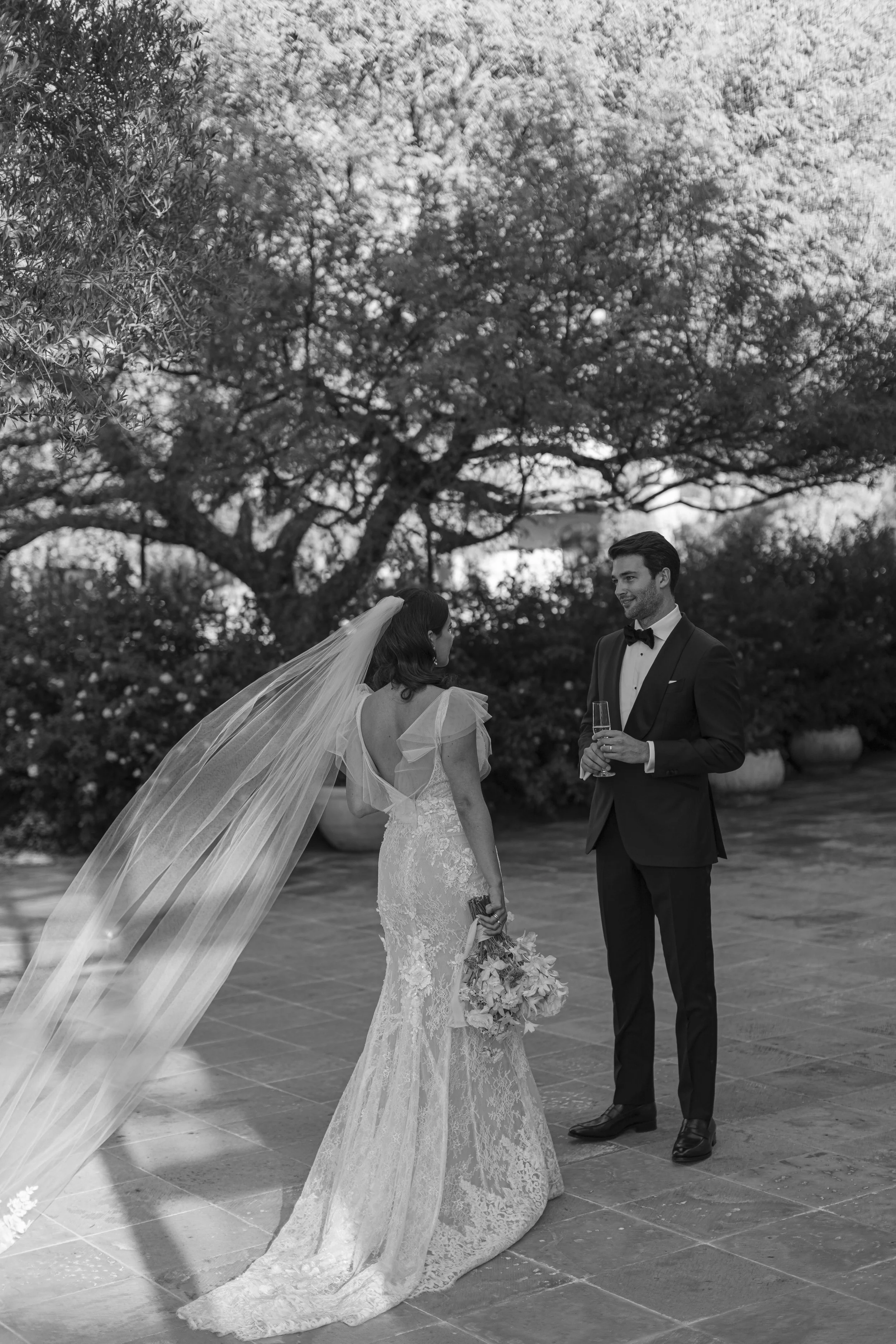 Una pareja de novios en una ceremonia nupcial al aire libre, con la novia con vestido de encaje y velo largo, y el novio con traje negro y pajarita, en un entorno con árboles y plantas.
