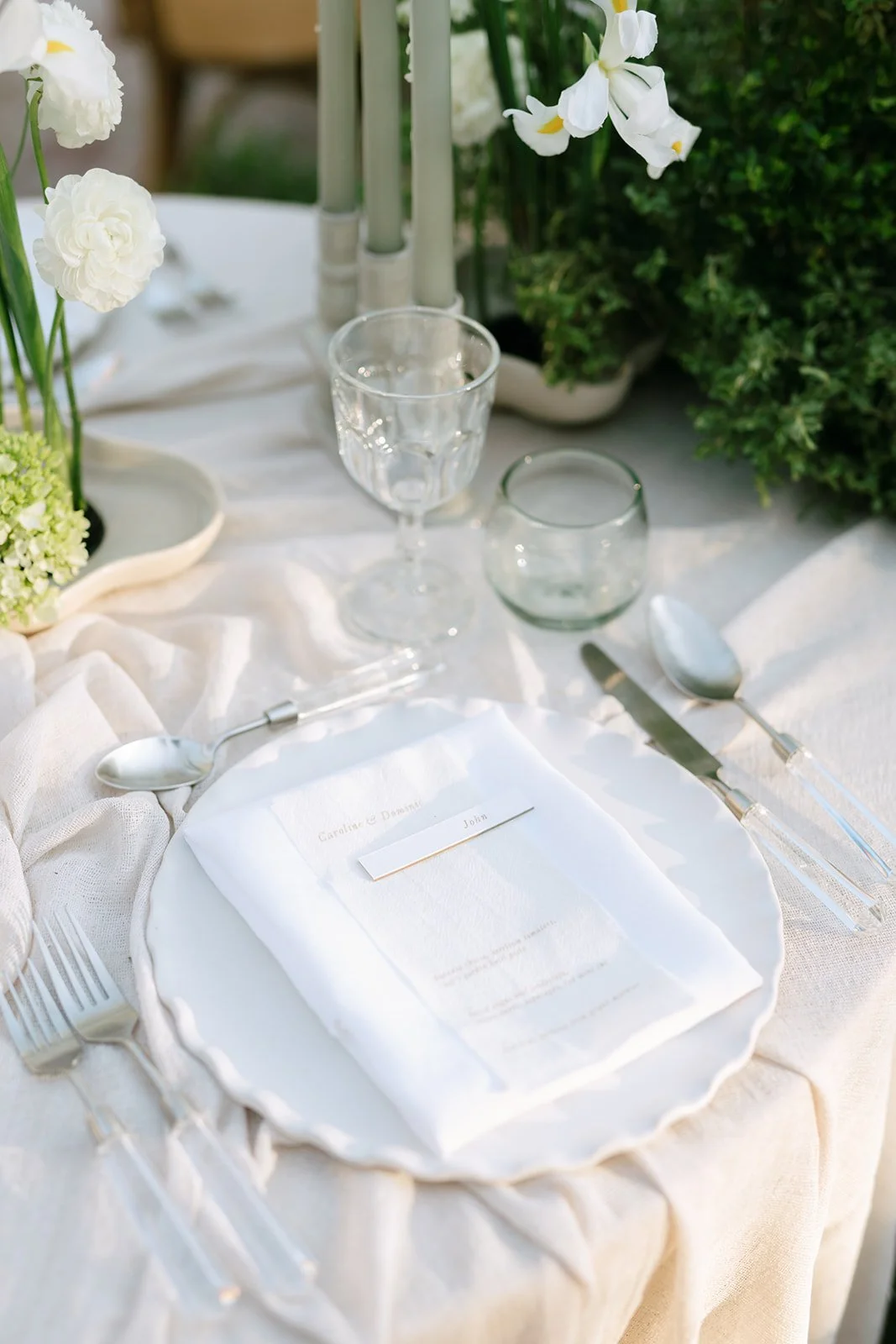 Mesa decorada con vajilla blanca y utensilios de plata, con un menú y un nombre, rodeada de flores blancas y plantas verdes en un ambiente luminoso.