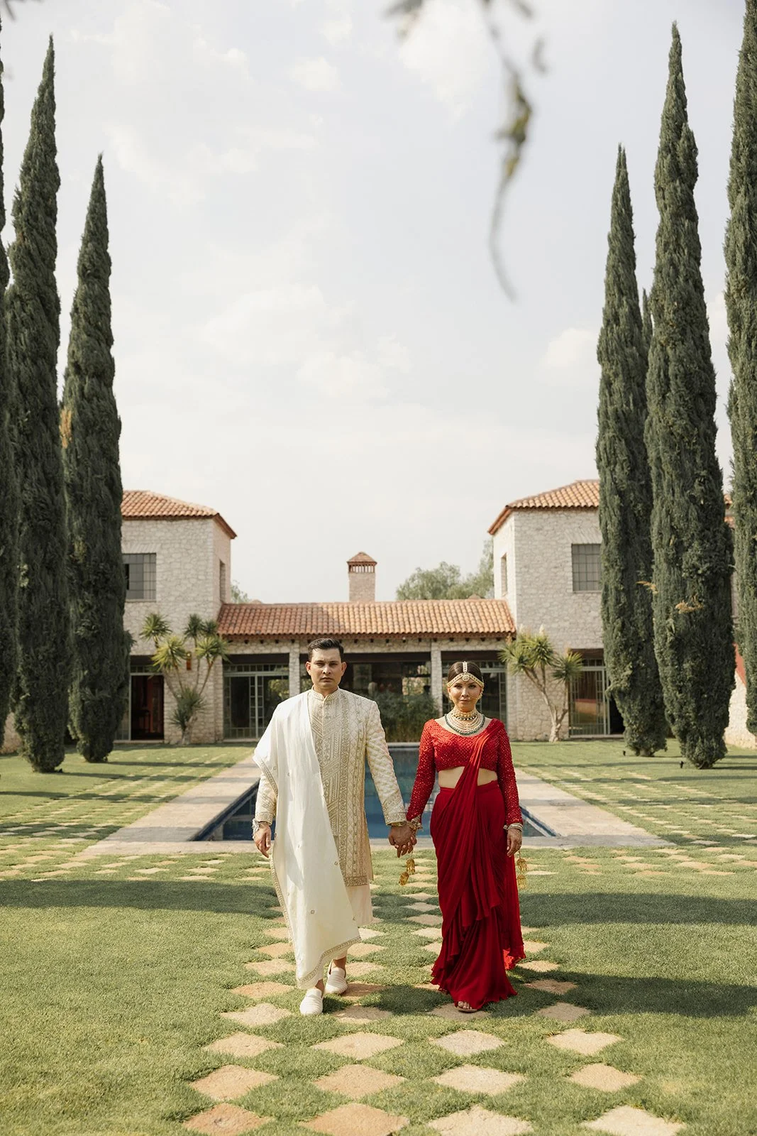 Pareja en vestimenta tradicional, caminando hacia la cámara en un jardín elegante con árboles altos y casa de fondo.
