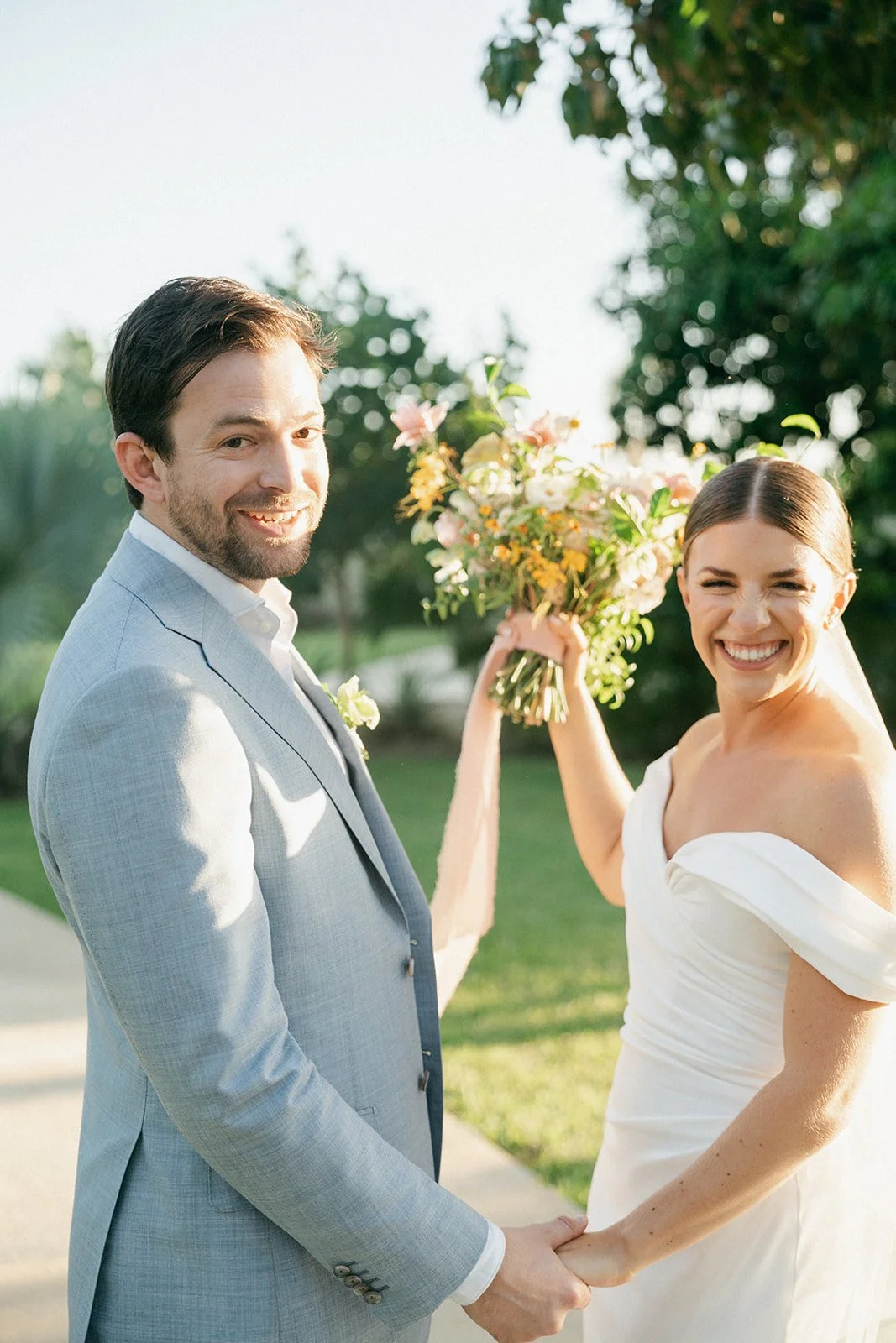 Pareja de novios en boda al aire libre, la novia con vestido blanco y el novio con saco gris, ambos sonriendo y sujetándose de las manos, la novia sostiene un ramo de flores.