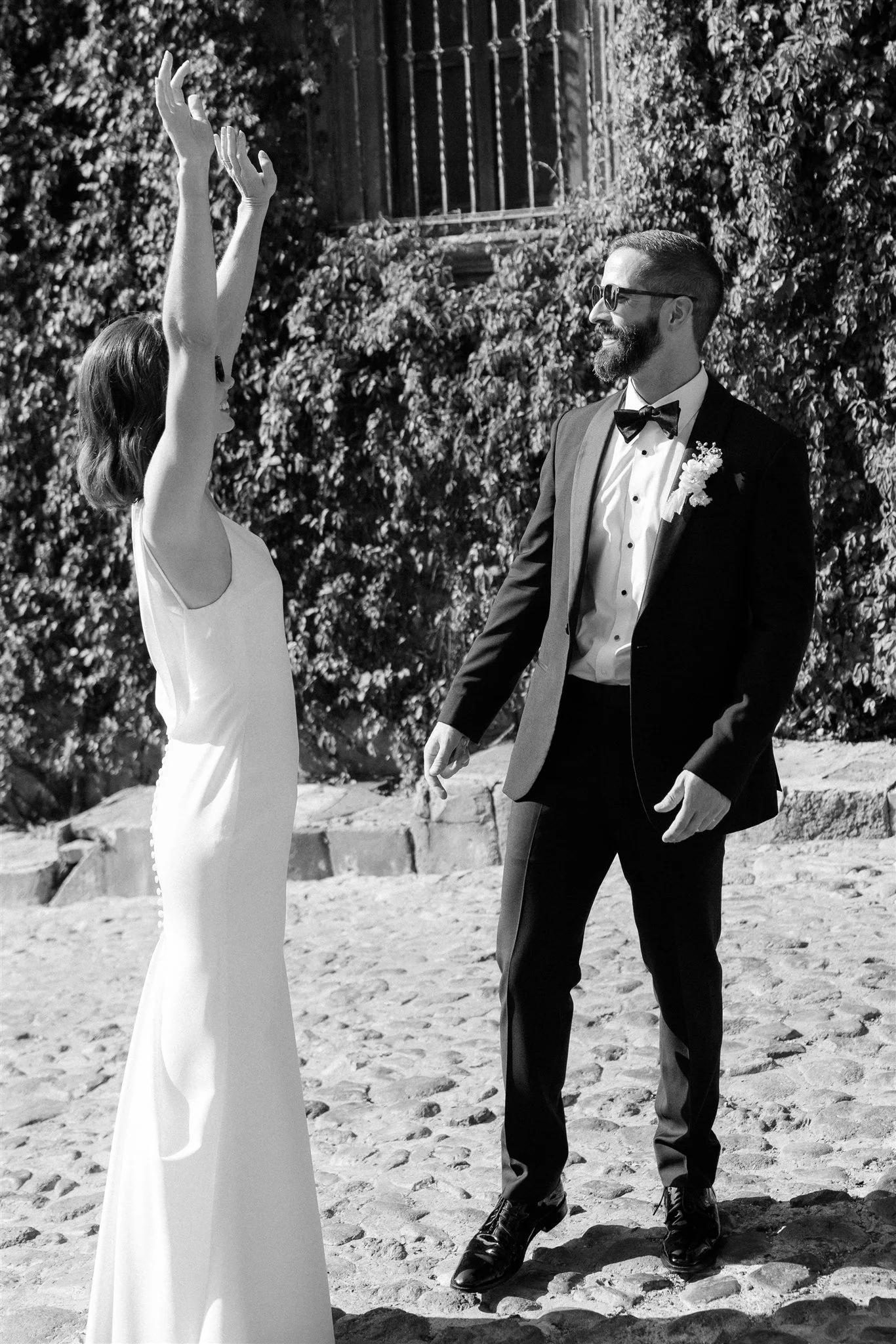 Una pareja casándose al aire libre, la mujer con vestido blanco y los brazos levantados y el hombre con traje negro y gafas, sonrientes.