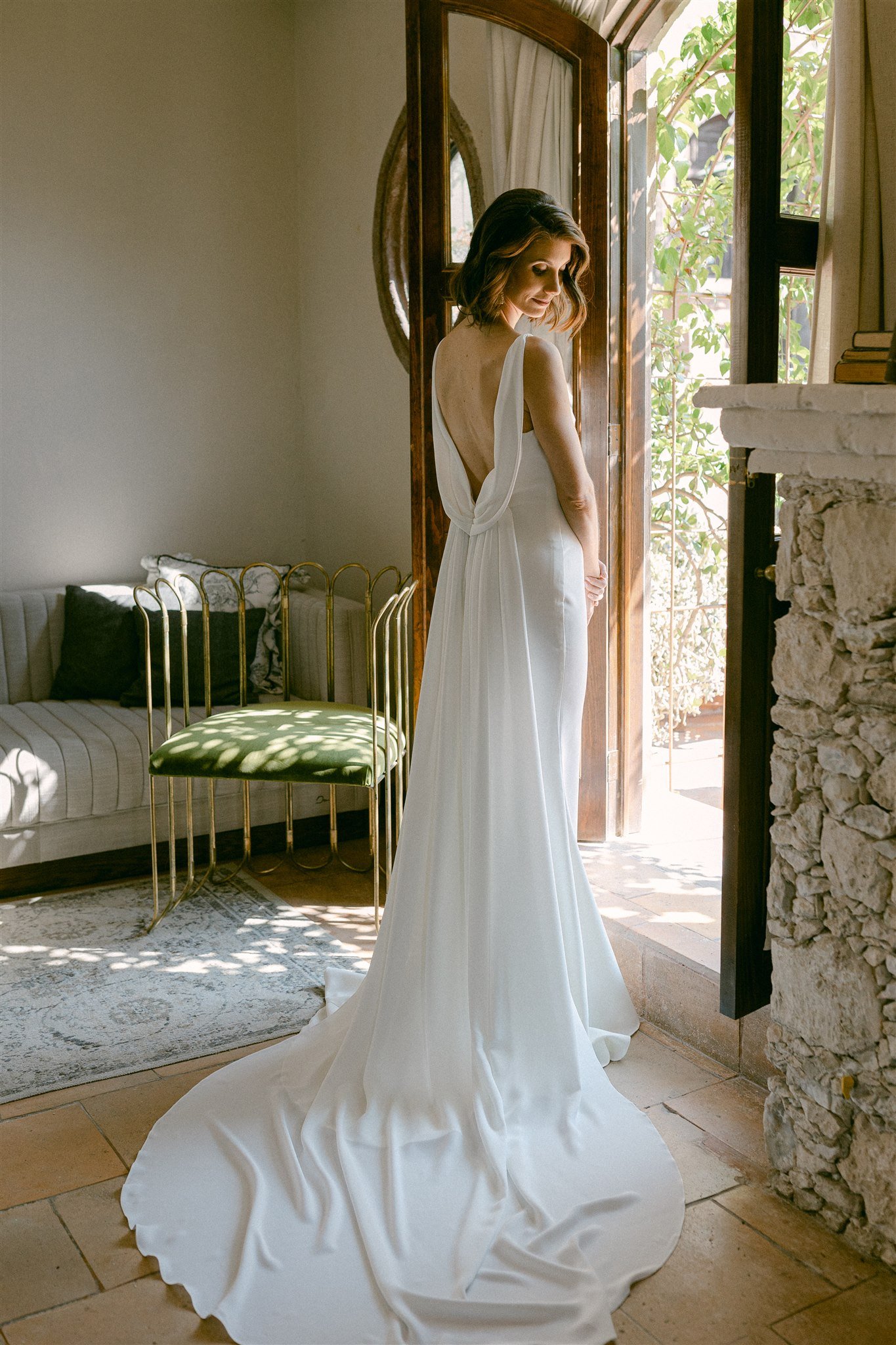 Mujer de espaldas con vestido de novia blanco, de manga larga y escote profundo en la espalda, frente a una puerta abierta que deja entrar luz natural y se ven plantas afuera.