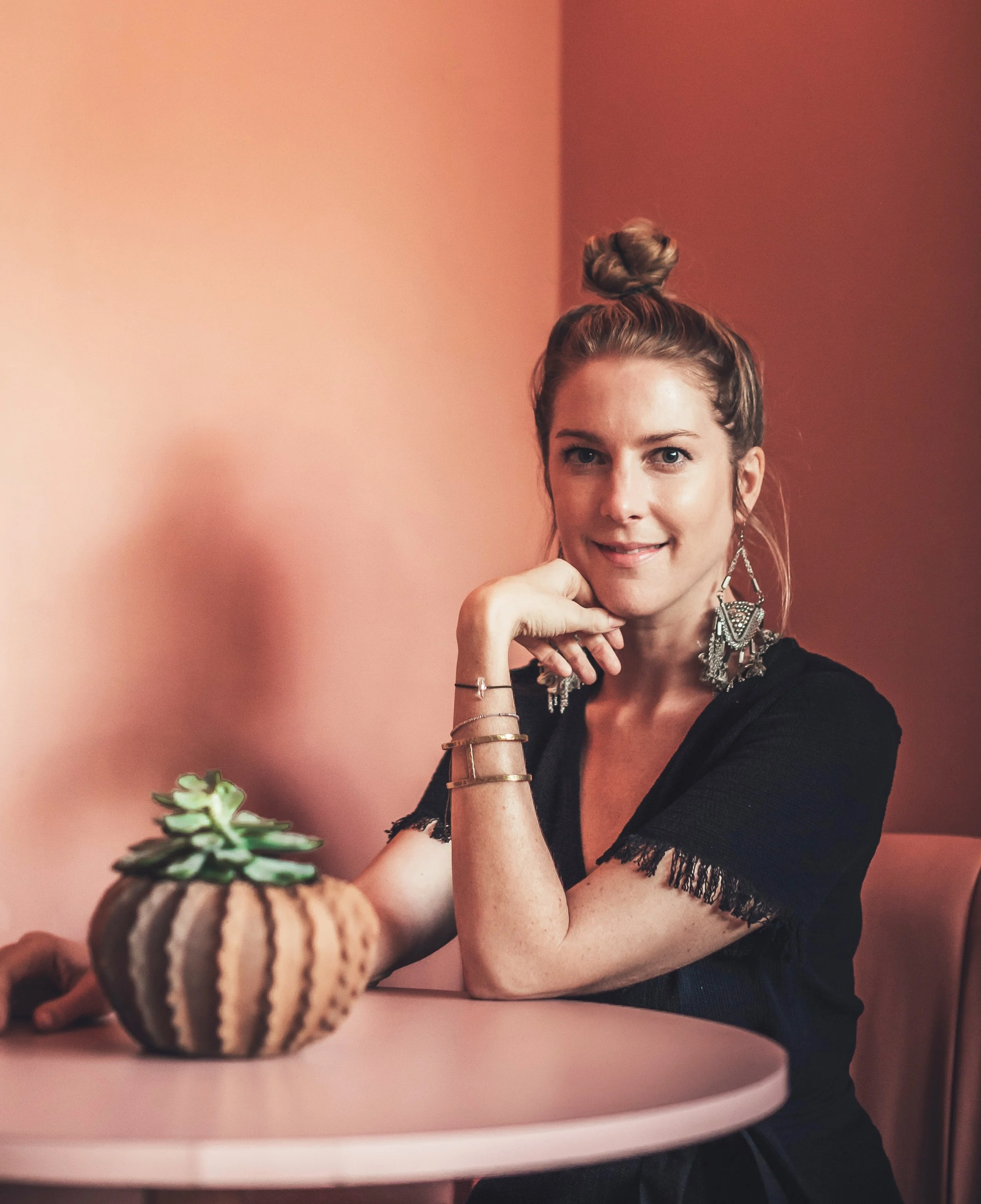 A woman with styled hair in a bun, wearing large earrings, sitting at a pink table with a small potted cactus plant, smiling at the camera.