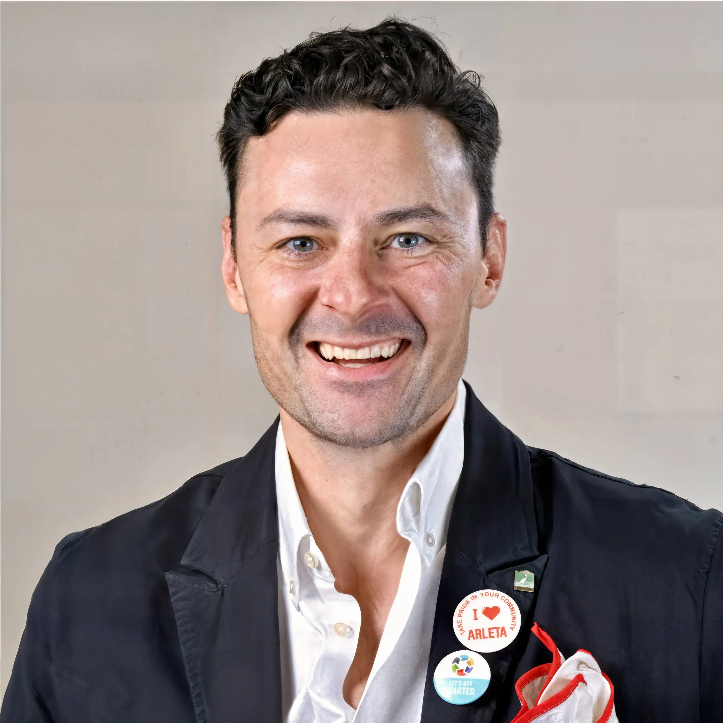 A smiling man with short dark hair, wearing a white shirt and a dark jacket with badges, including one that says 'I love Arleta' and another of a recycling symbol, standing in front of a neutral background.