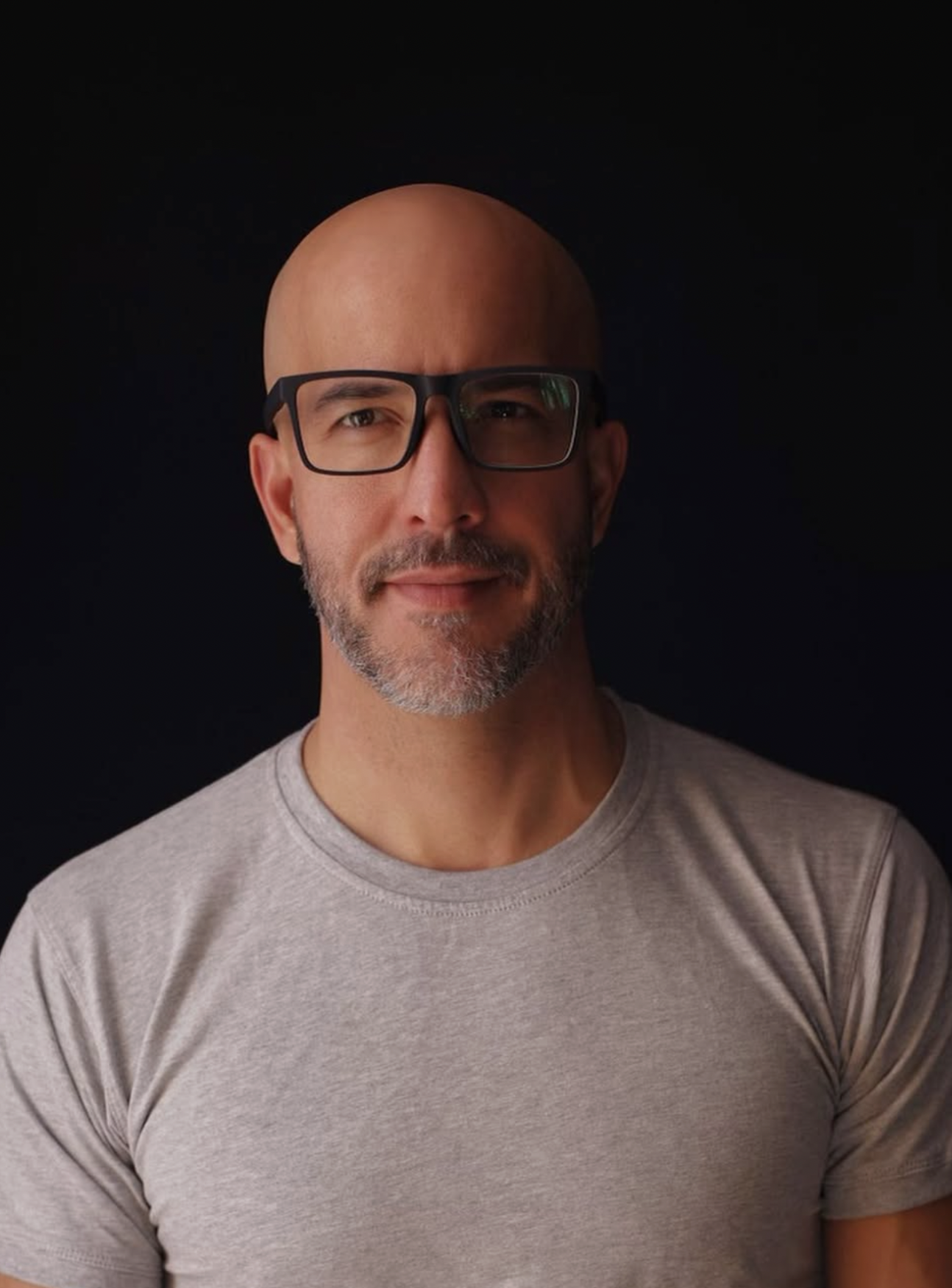 A middle-aged man with a bald head, glasses, and a short beard, wearing a light gray t-shirt, standing against a dark background.