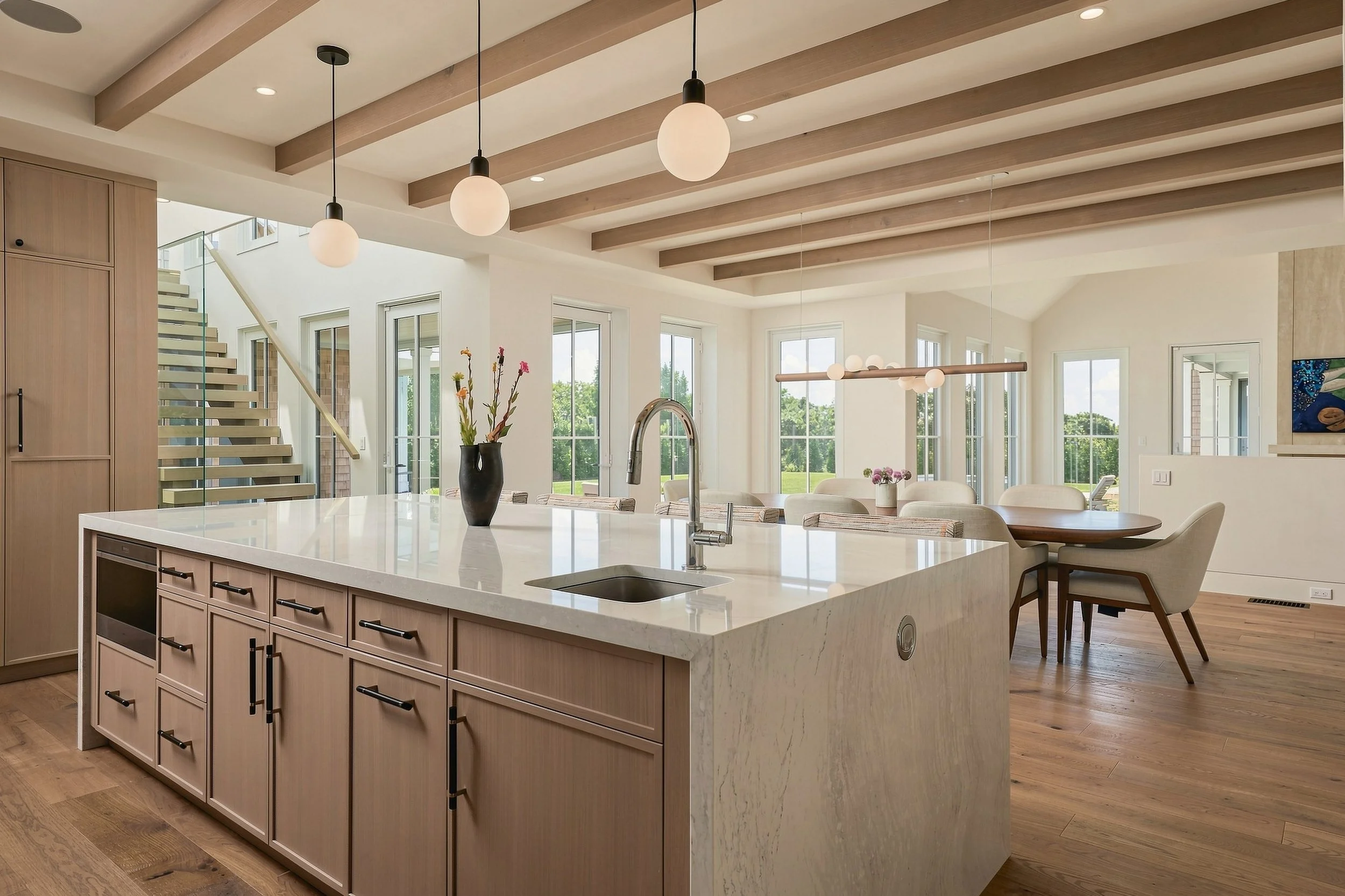 Open kitchen and dining area with a waterfall island, light wood cabinetry, exposed beams and globe pendants