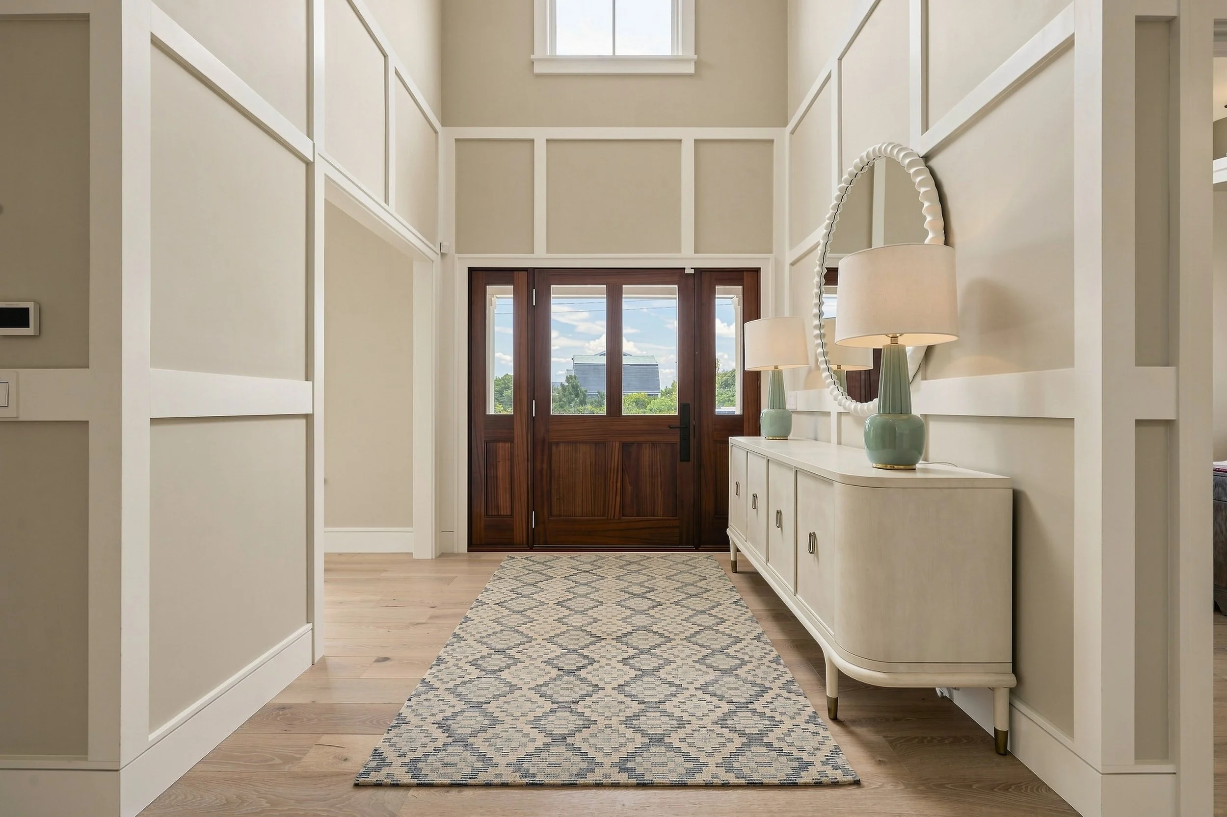 Double-height entry foyer with paneled walls, a wood front door, patterned runner and mirrored console