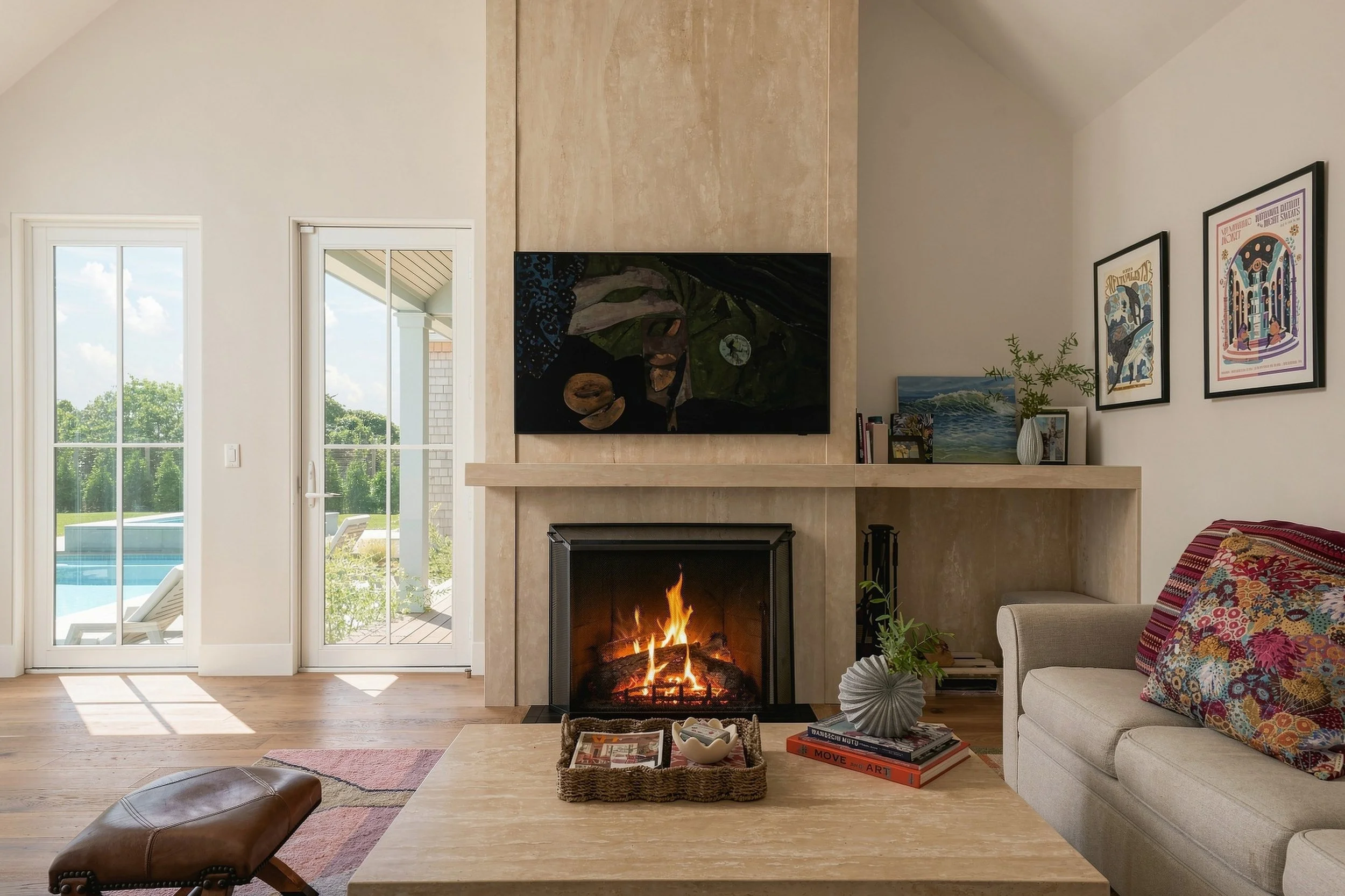 Living room fireplace with a pale stone surround, built-in shelf, colorful art and views of the pool