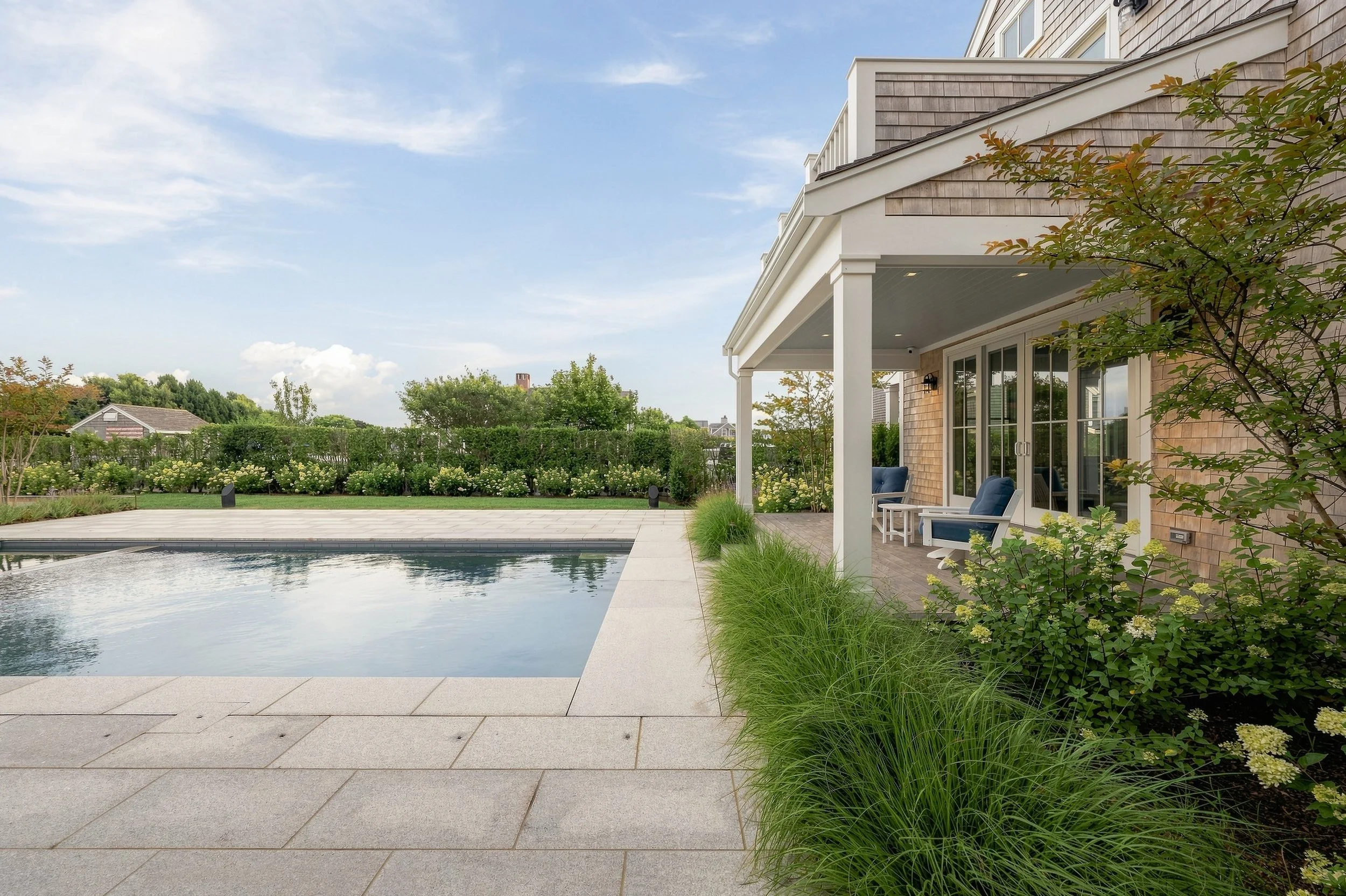 Poolside view of a covered porch with blue chairs, hydrangeas and cedar-shingle siding
