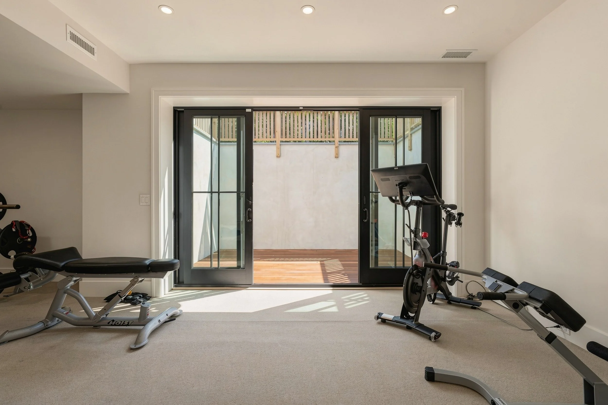 Home gym with a stationary bike, rowing machine and weight bench facing black-framed patio doors