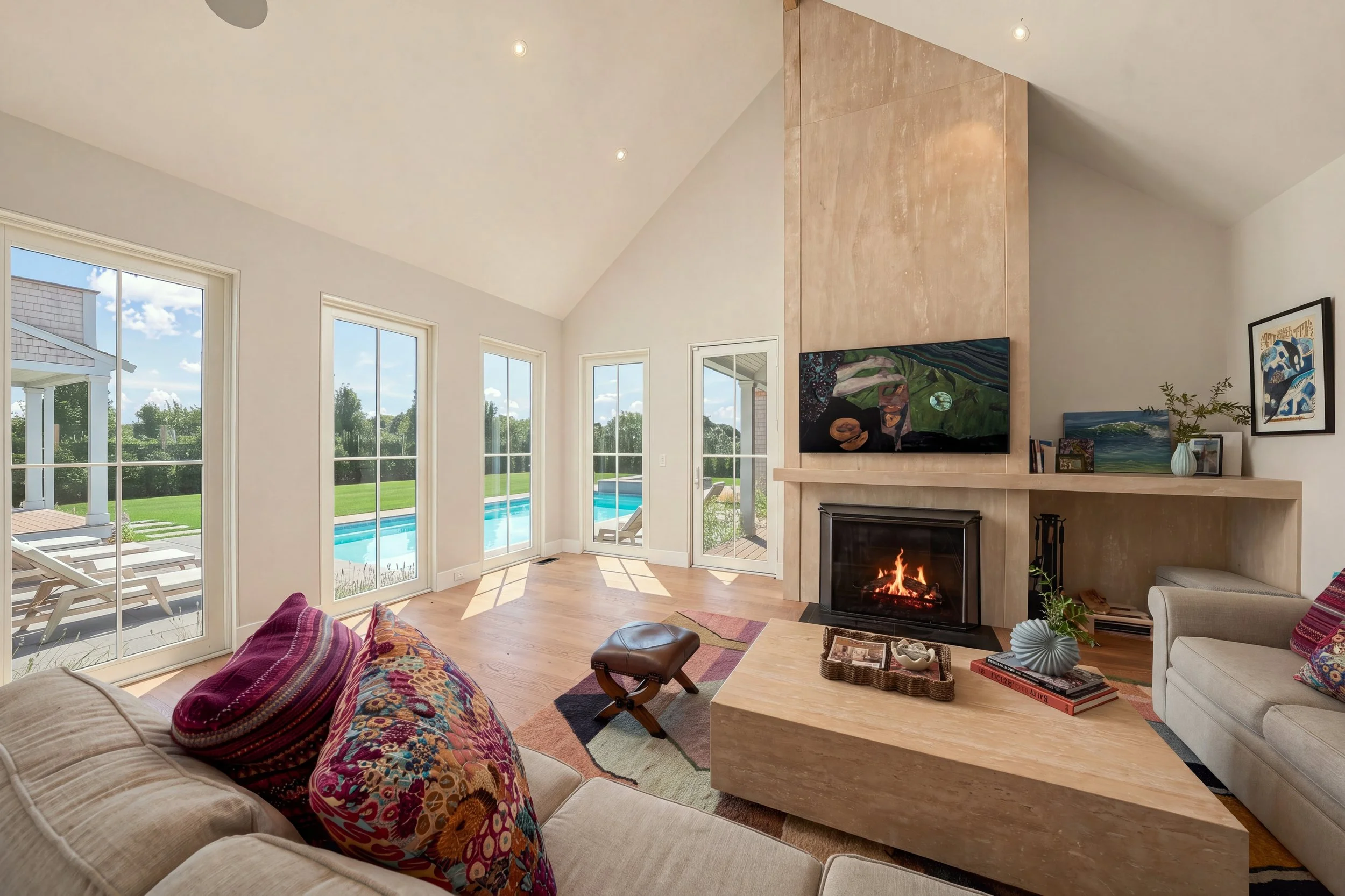 Vaulted living room with a pale stone fireplace, colorful textiles and tall windows overlooking the pool
