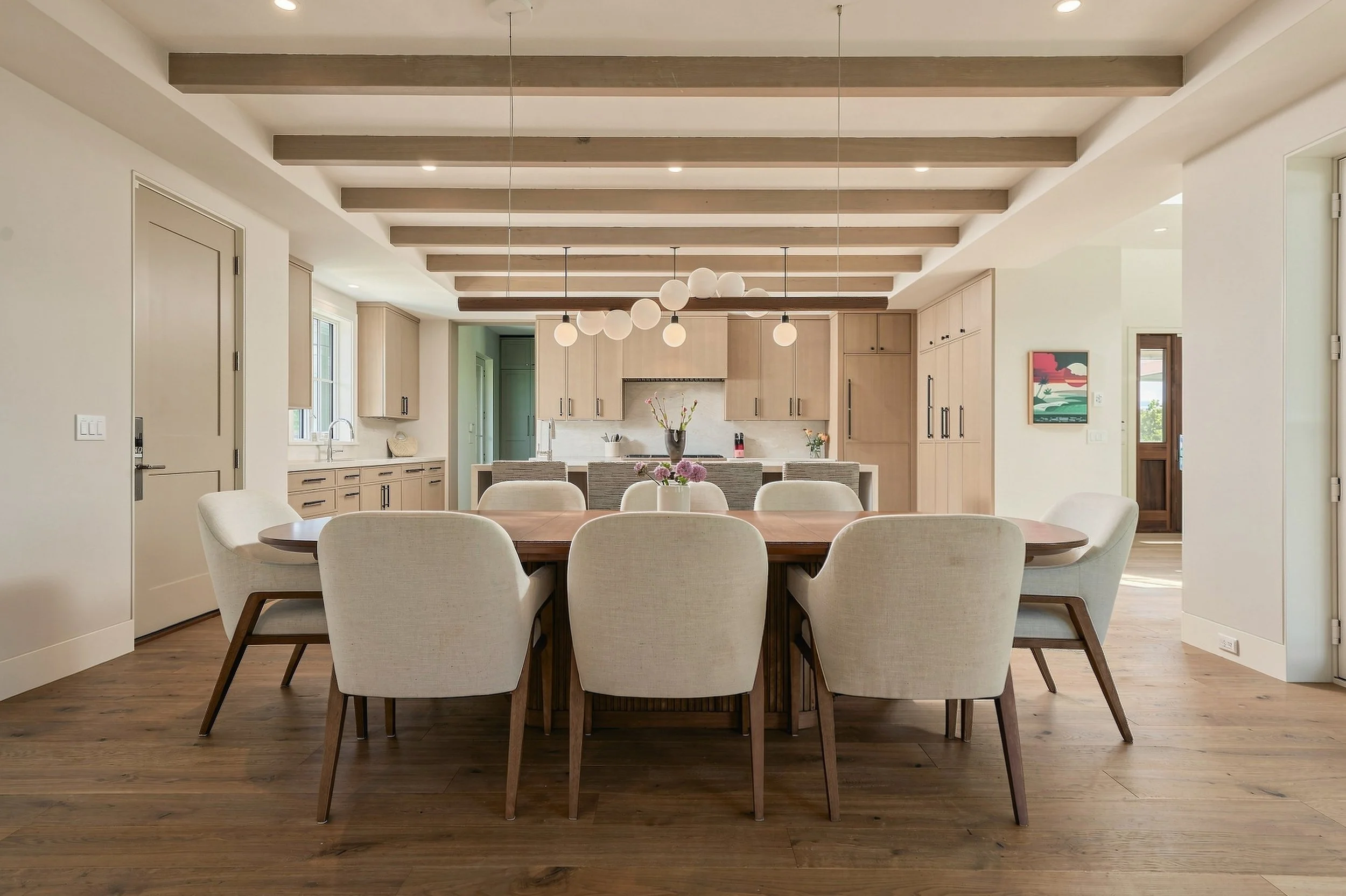 Dining room with an oval wood table and upholstered chairs beneath globe pendants, opening to the kitchen