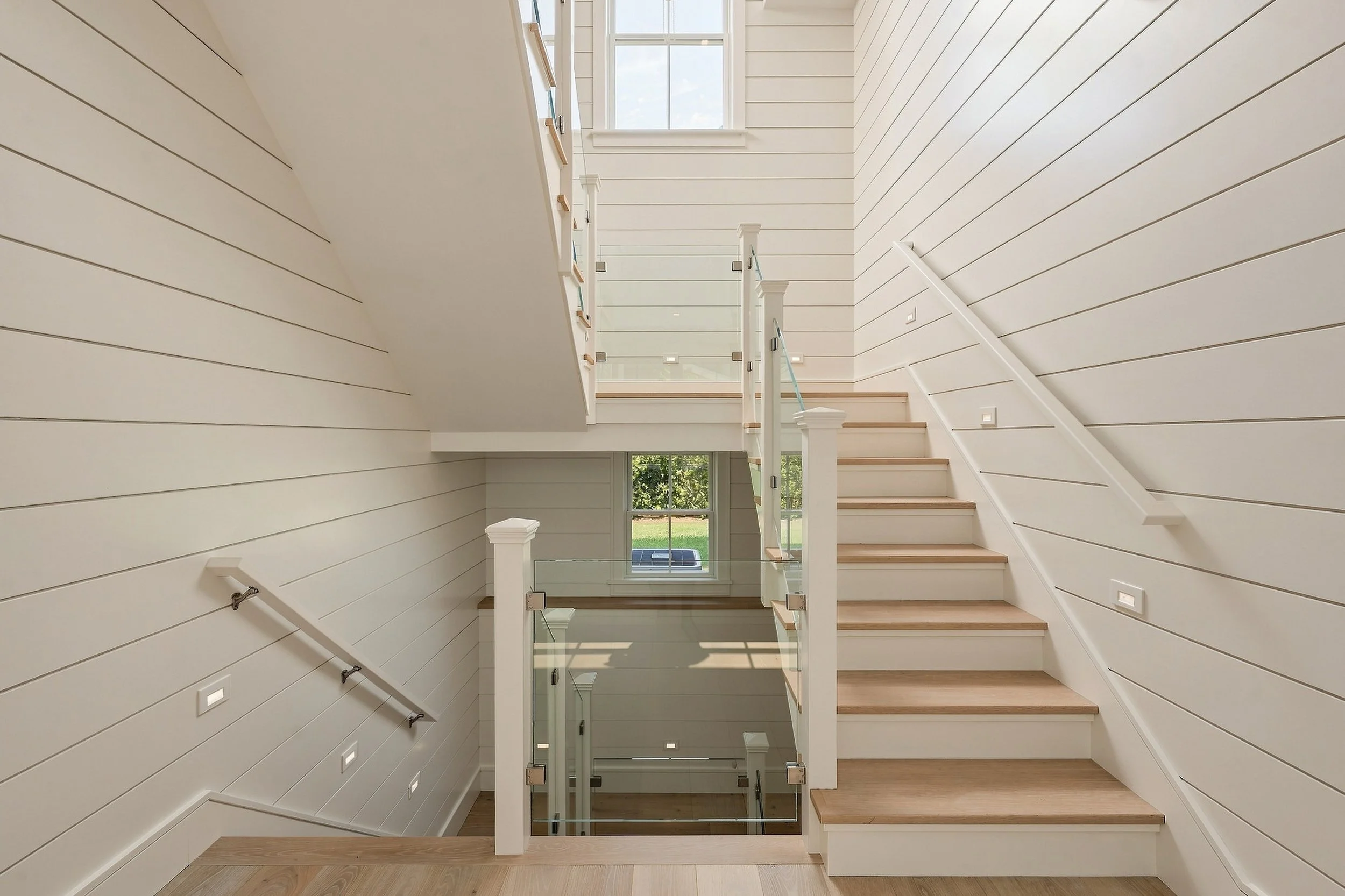 Three-story stair hall with white shiplap walls, oak treads, glass railings and tall windows