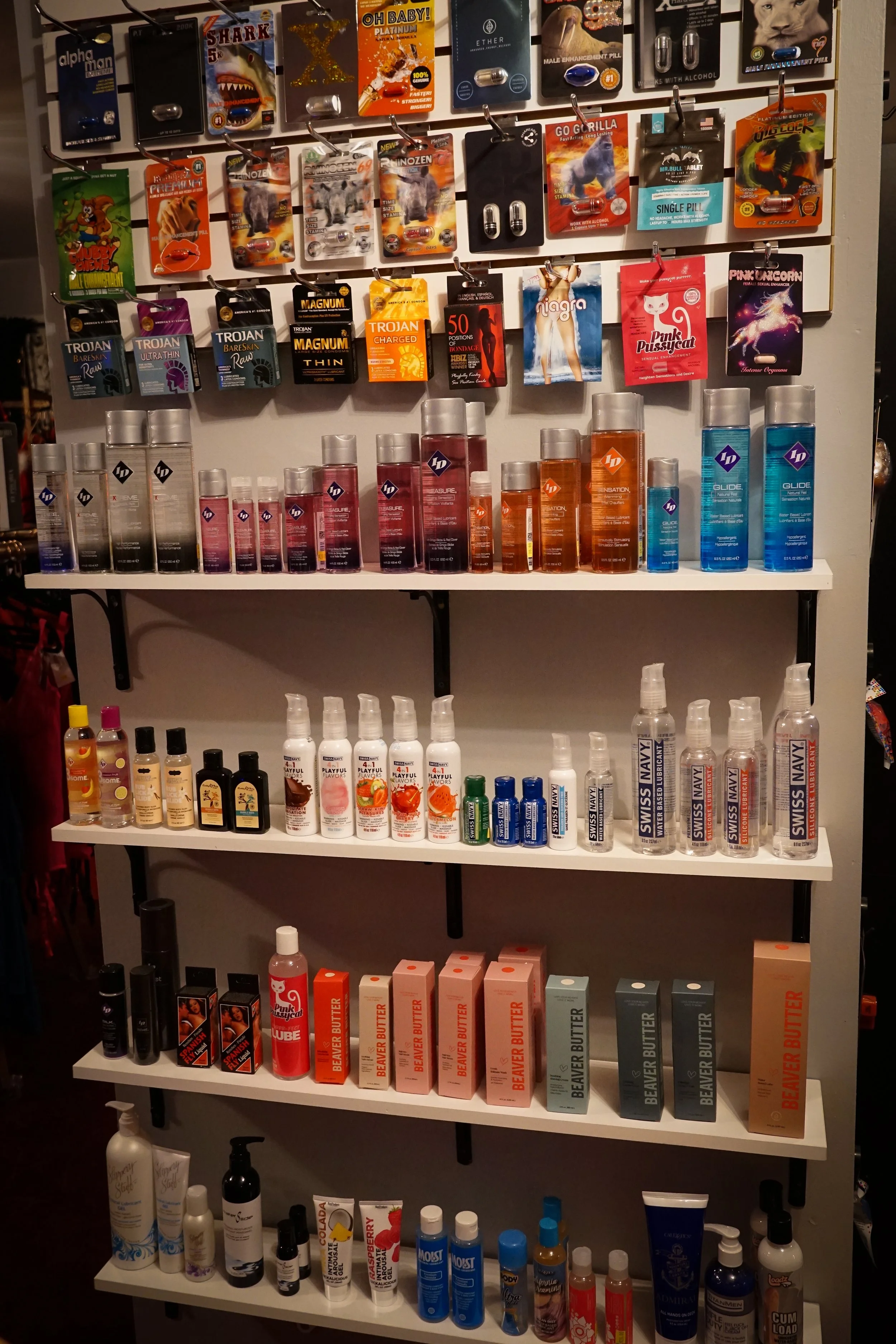 Display of various personal lubricants, massage oils, and adult products arranged on white shelves with colorful packaging and bottles.