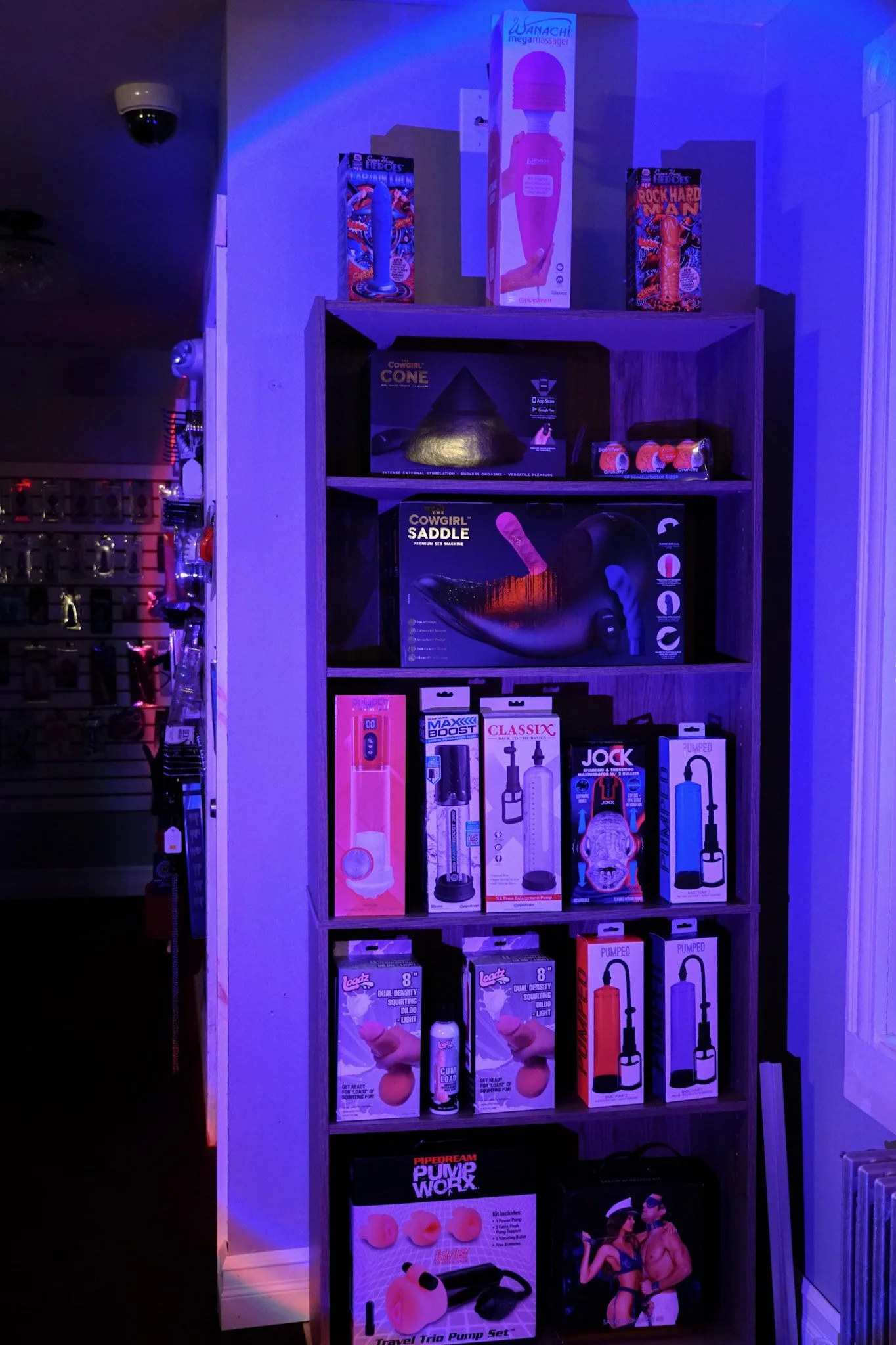 Shelves displaying adult toys and products, including vibrators, pumps, and other intimate accessories, illuminated by purple-blue lighting.