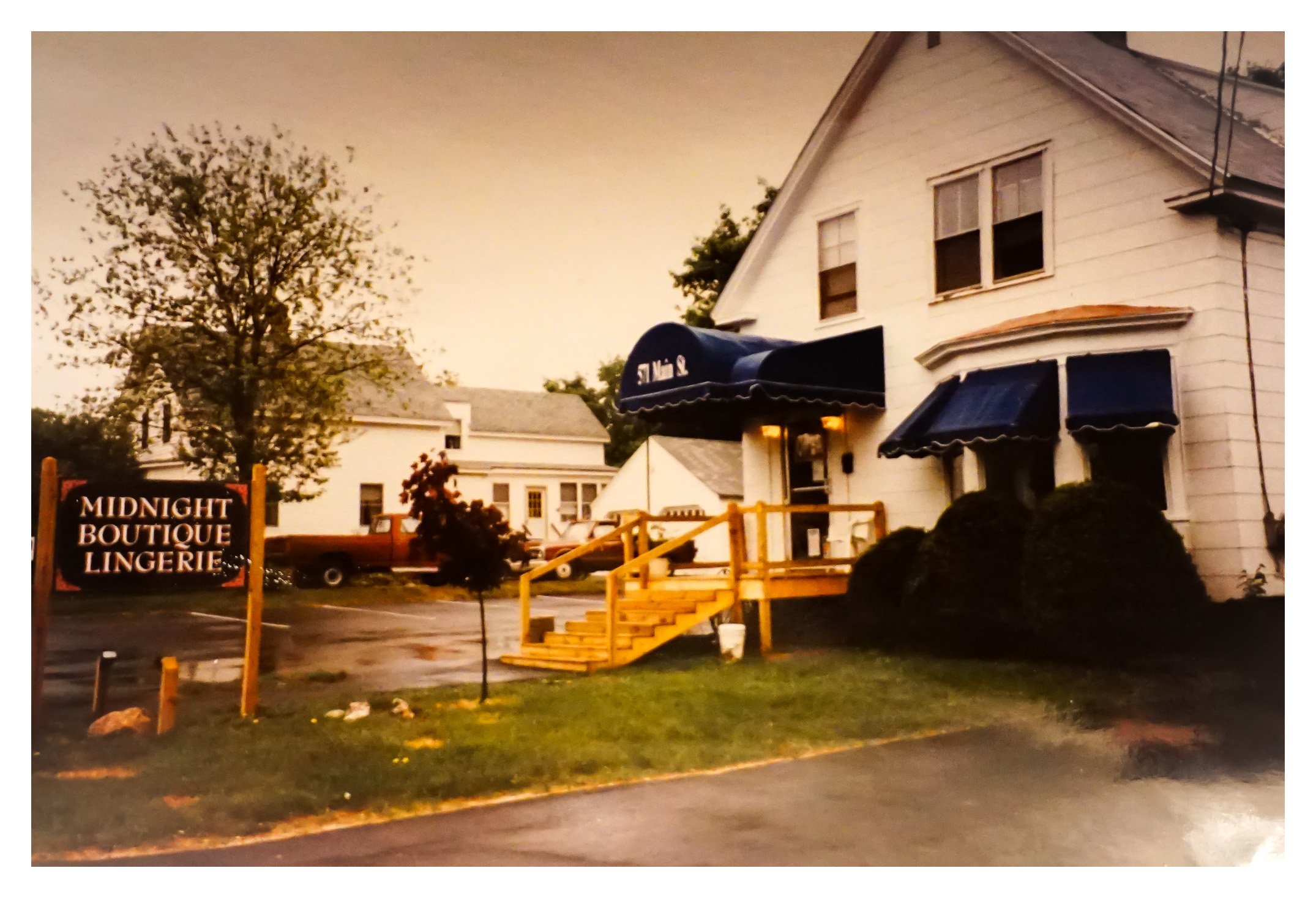 A white building with blue awnings and a wooden staircase leading to a door. A sign in front reads 'Midnight Boutique Lingerie'. There are cars parked in the background, some trees, and the scene appears to be during early evening or dusk.