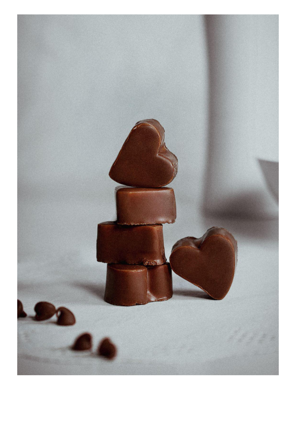 Stack of four chocolates shaped like hearts, with additional hearts in the background, on a white surface.